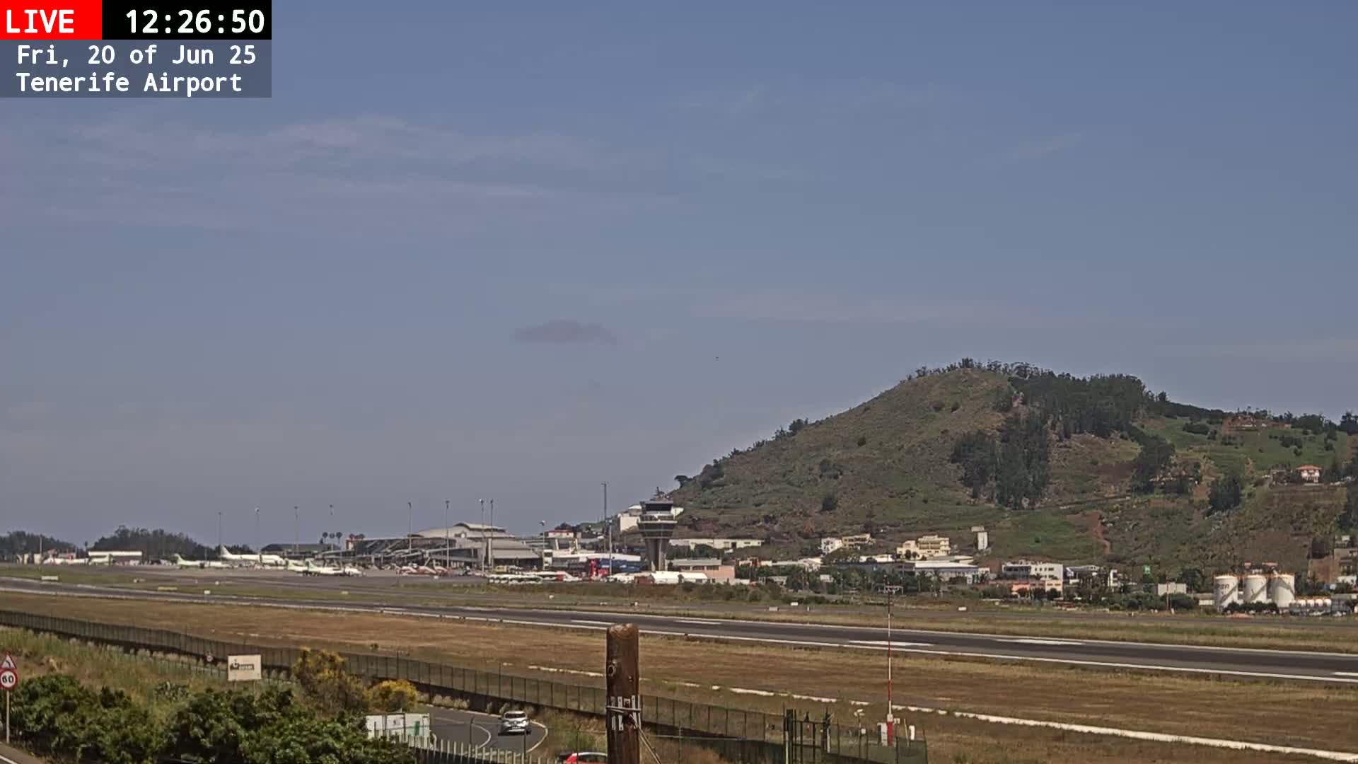 An airport runway with several airplanes parked near the terminal buildings, a control tower, and a large green hill in the background under a mostly clear sky.
