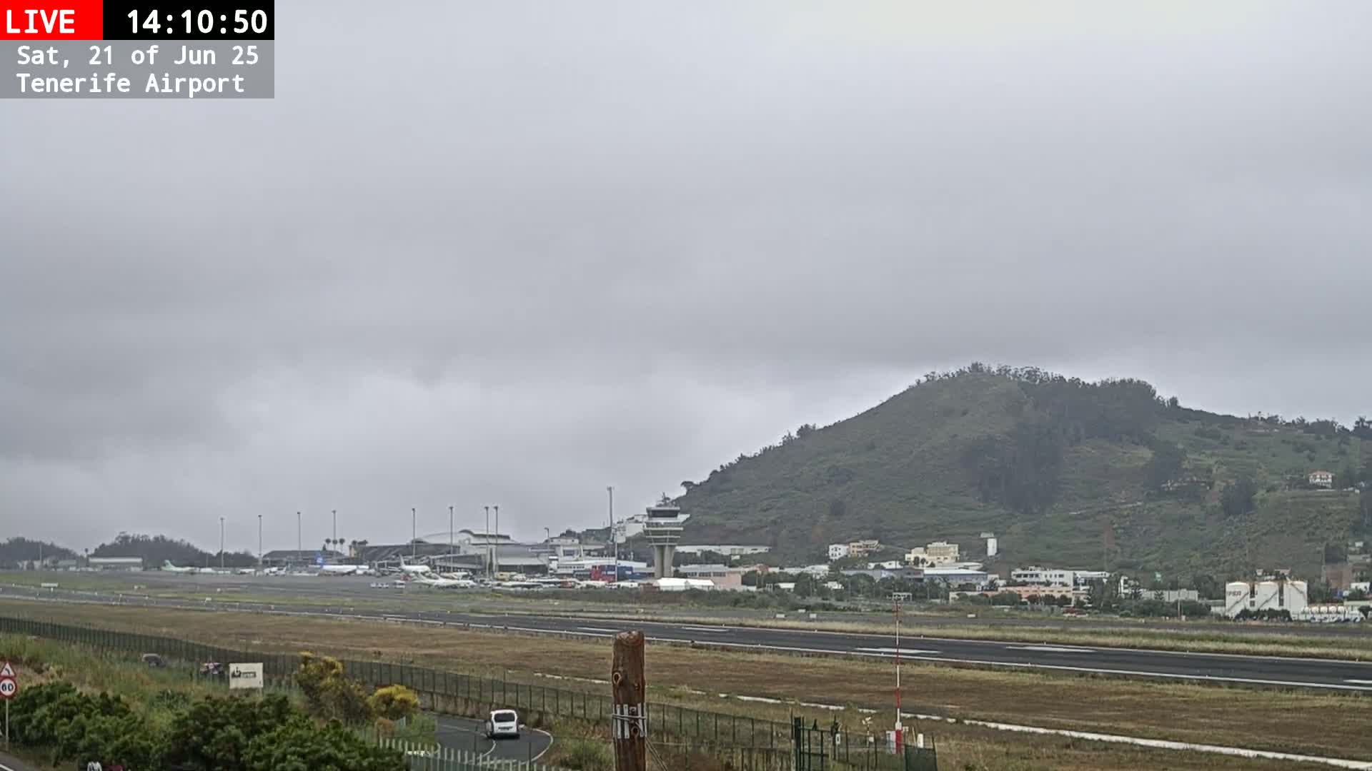 A view of an airport runway with several airplanes, surrounded by buildings and a green hill under an overcast sky.