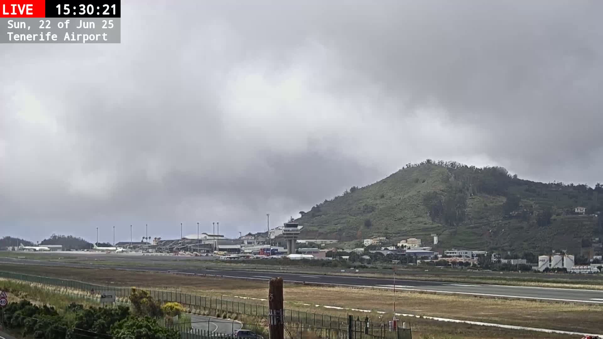 An airport runway and terminal are visible beneath a cloudy sky, with a large hill in the background.