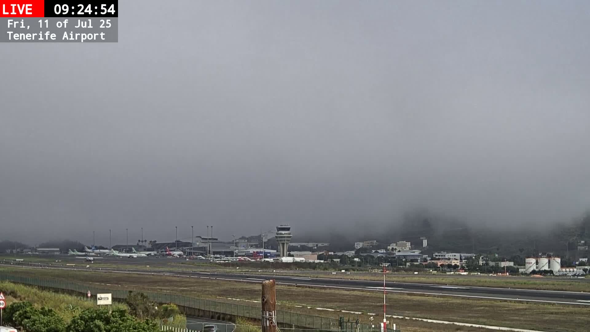 A foggy view of an airport runway with several airplanes parked and a control tower visible in the distance.