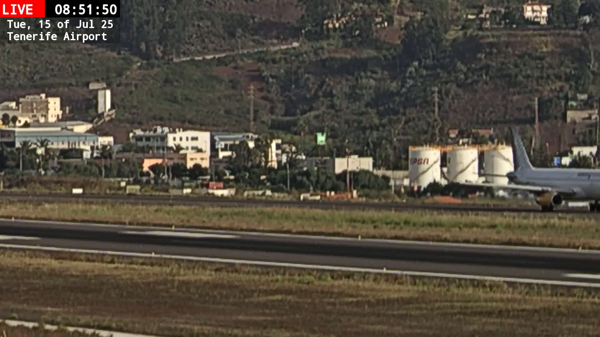 A passenger airplane taxis on an airport runway past several buildings and large fuel tanks, under a mostly clear sky.