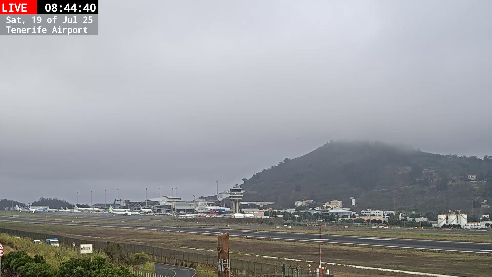 A foggy view of an airport runway with several airplanes parked, a control tower, and a hill shrouded in mist in the background.