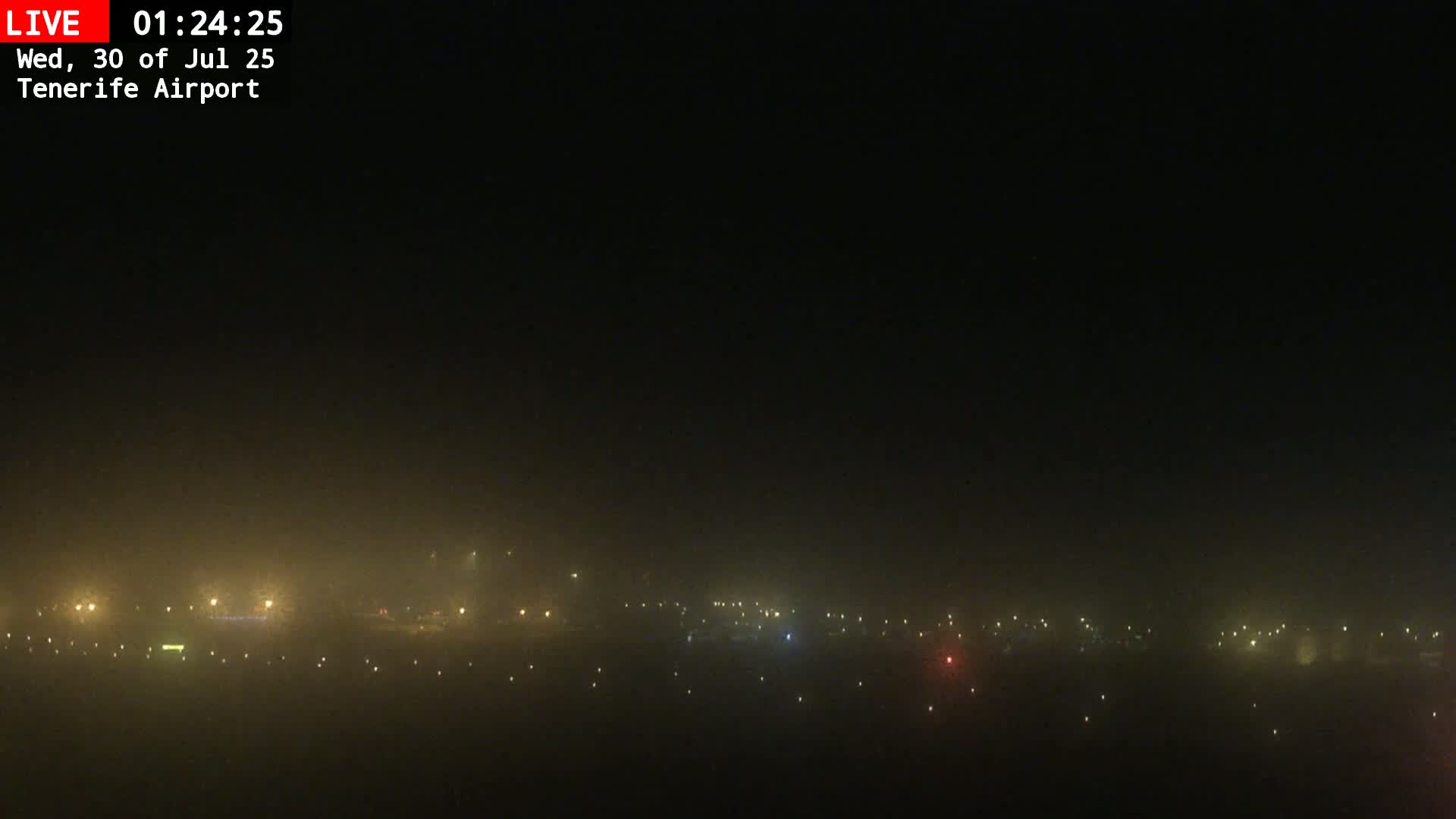 A foggy view of an airport runway with several airplanes parked, a control tower, and a hill shrouded in mist in the background.