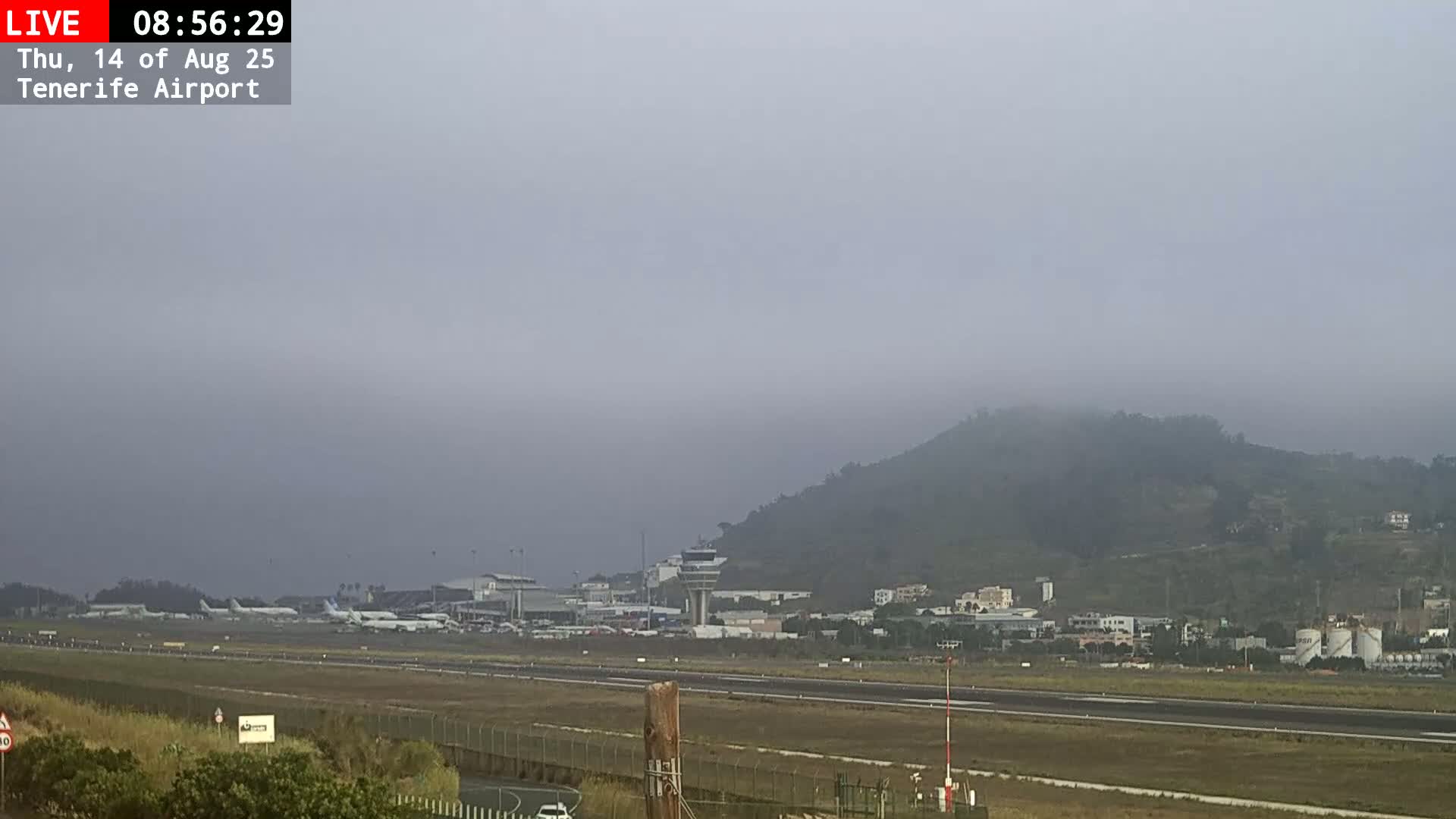A foggy view of an airport runway, terminal buildings, and airplanes, with a hill shrouded in mist in the background.