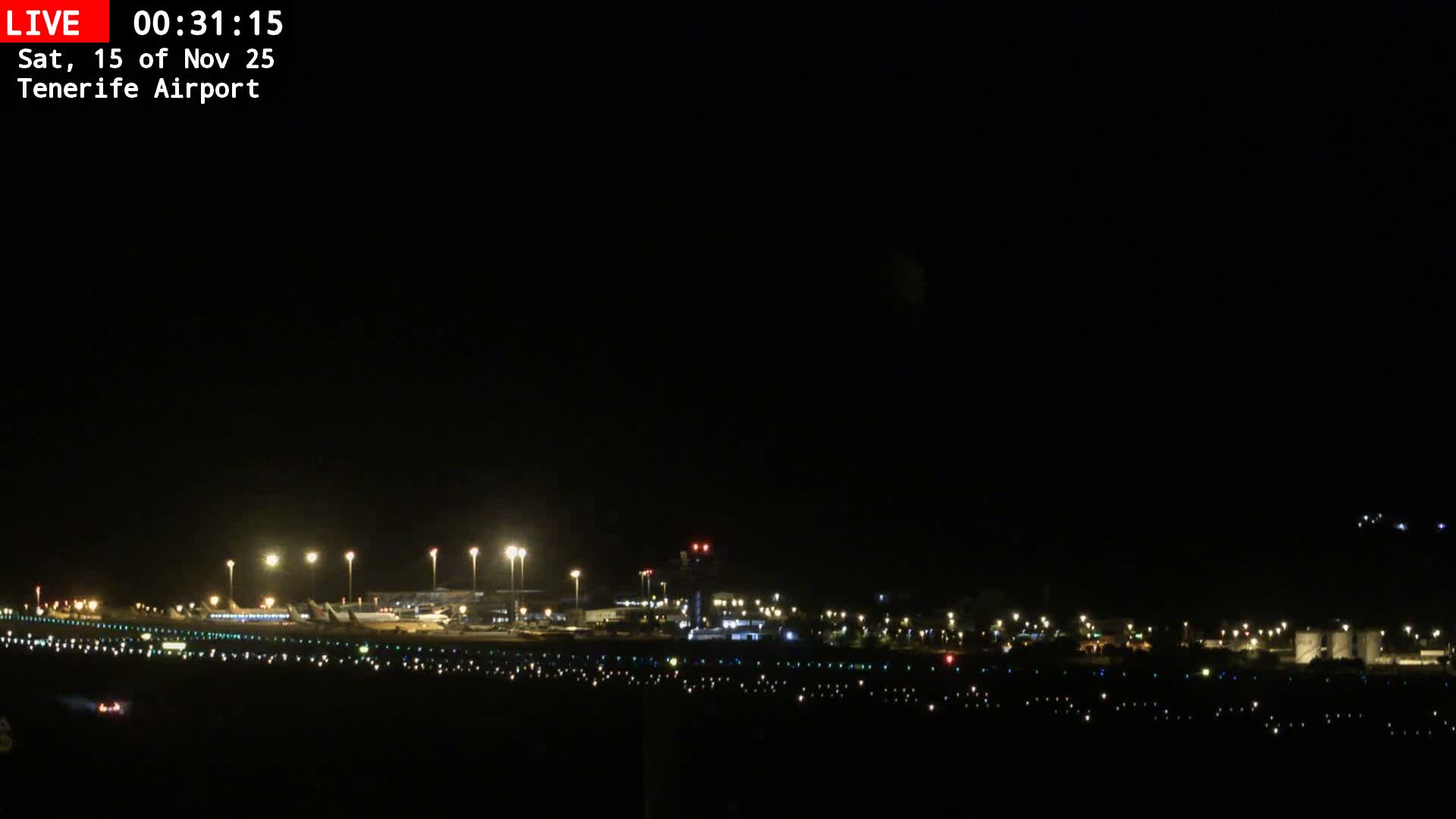 A clear, dark night sky hangs over an airport, where runways, taxiways, and terminal buildings are brightly illuminated, with several aircraft visible on the tarmac.