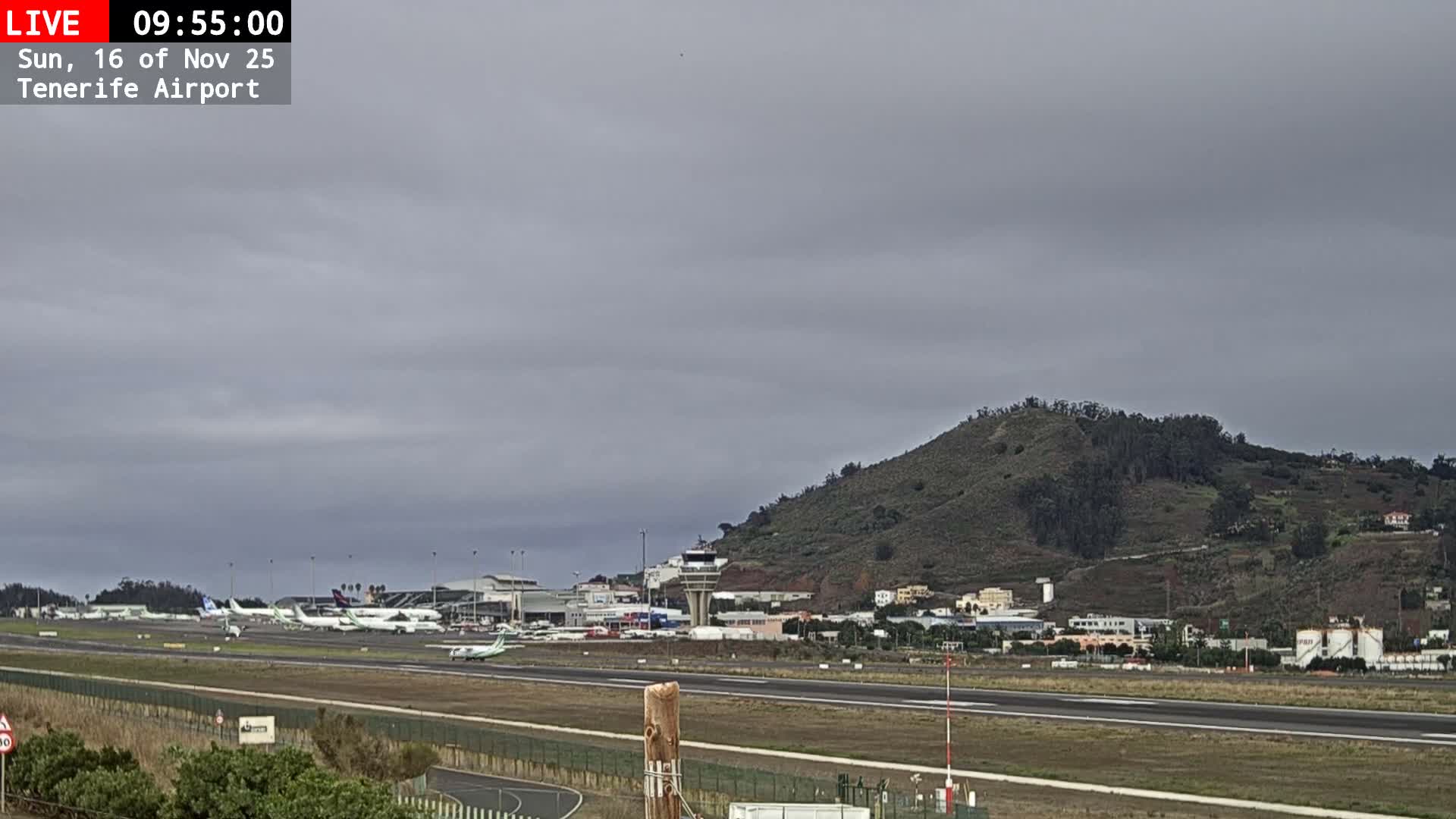 An overcast and cloudy sky hangs over an airport bustling with parked airplanes and buildings, set against a backdrop of a large, green-covered hill with scattered structures.