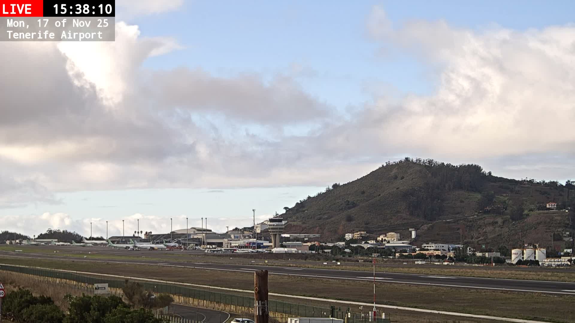 An outdoor wide shot reveals an airport with several aircraft on the tarmac and a control tower, backed by a large green and brown hill dotted with buildings, under a partly cloudy sky.