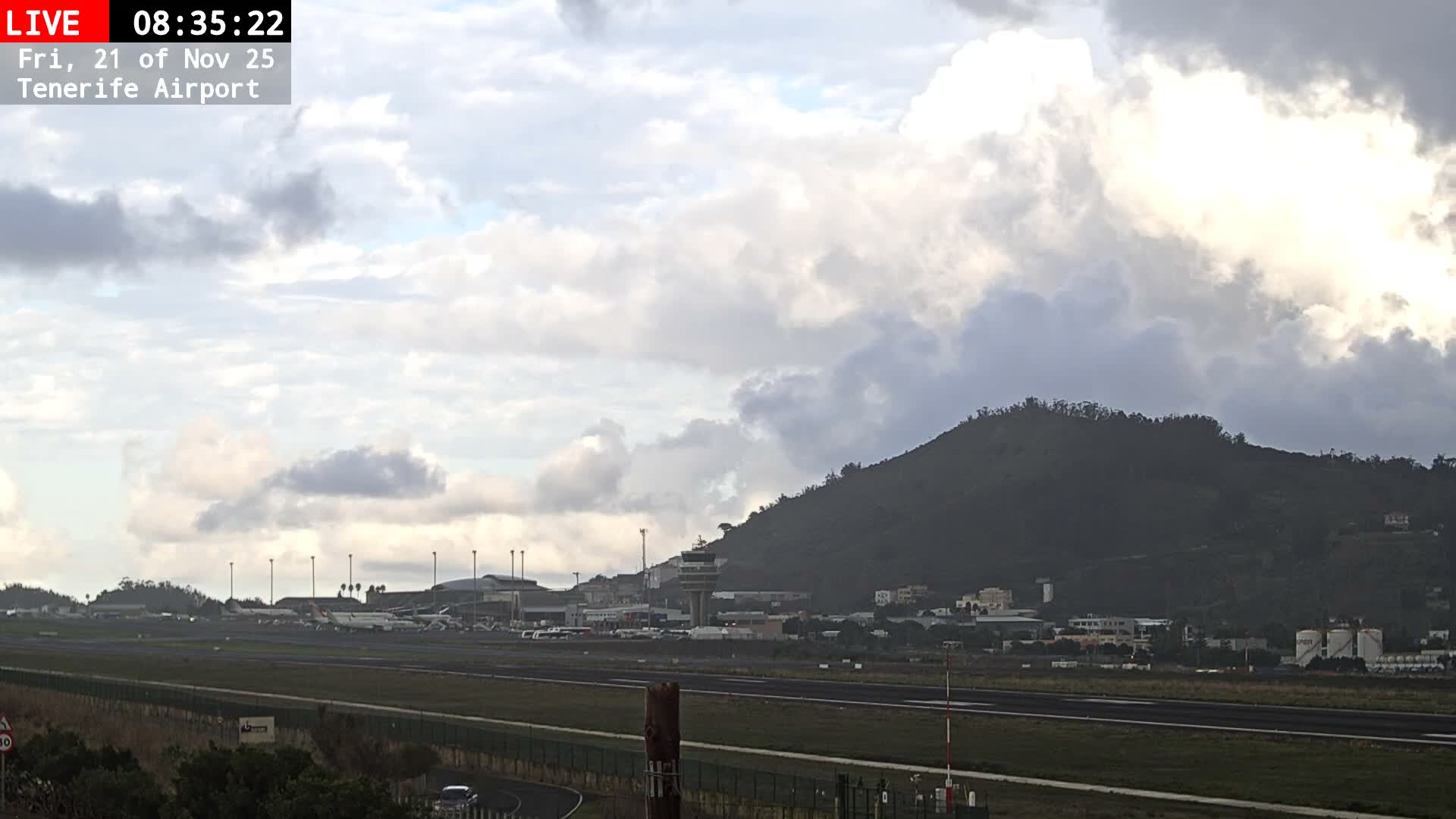 An airport runway with several parked airplanes and terminal buildings is visible under a partly cloudy sky, with a large, verdant mountain rising in the background dotted with scattered buildings.