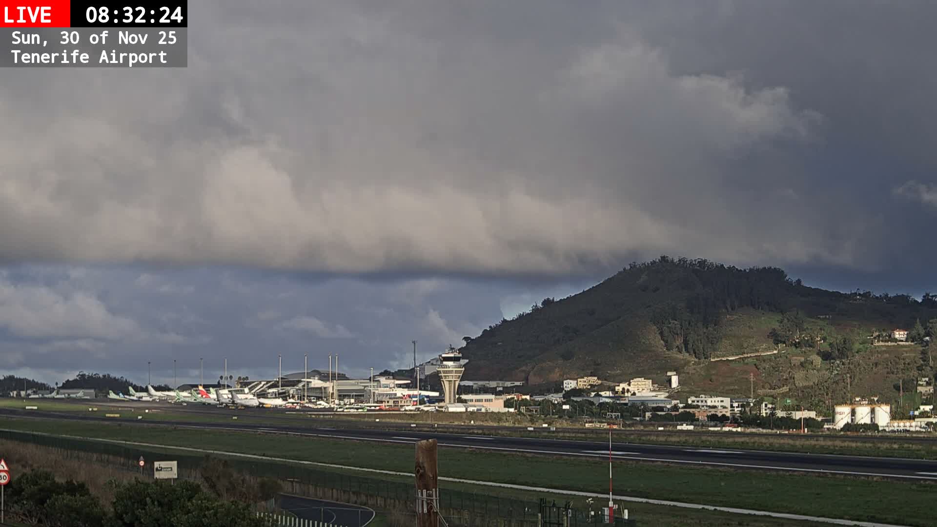 A wide shot of an airport reveals multiple parked airplanes, a terminal building, and a control tower, all set against a large, tree-covered mountain under a dramatically overcast sky with dark, textured clouds.