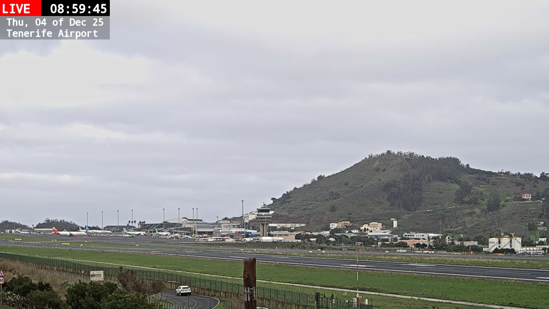 The image captures an overcast daytime view of an airport with multiple planes on the tarmac, a control tower, and terminal buildings, all set against a backdrop of green, tree-covered hills with scattered houses.