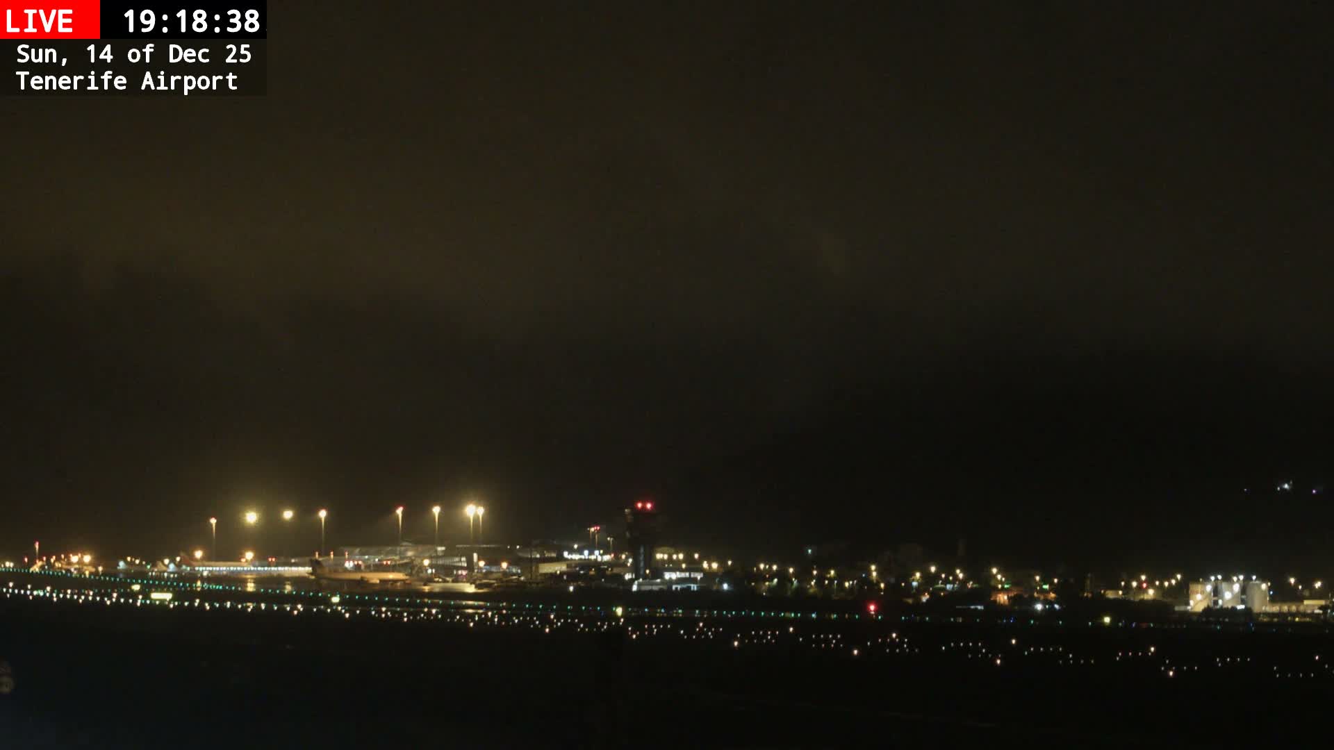 An airport runway and tarmac, featuring multiple airplanes, terminal buildings, and a control tower, are set against a backdrop of a large, tree-covered mountain under a uniformly overcast sky.