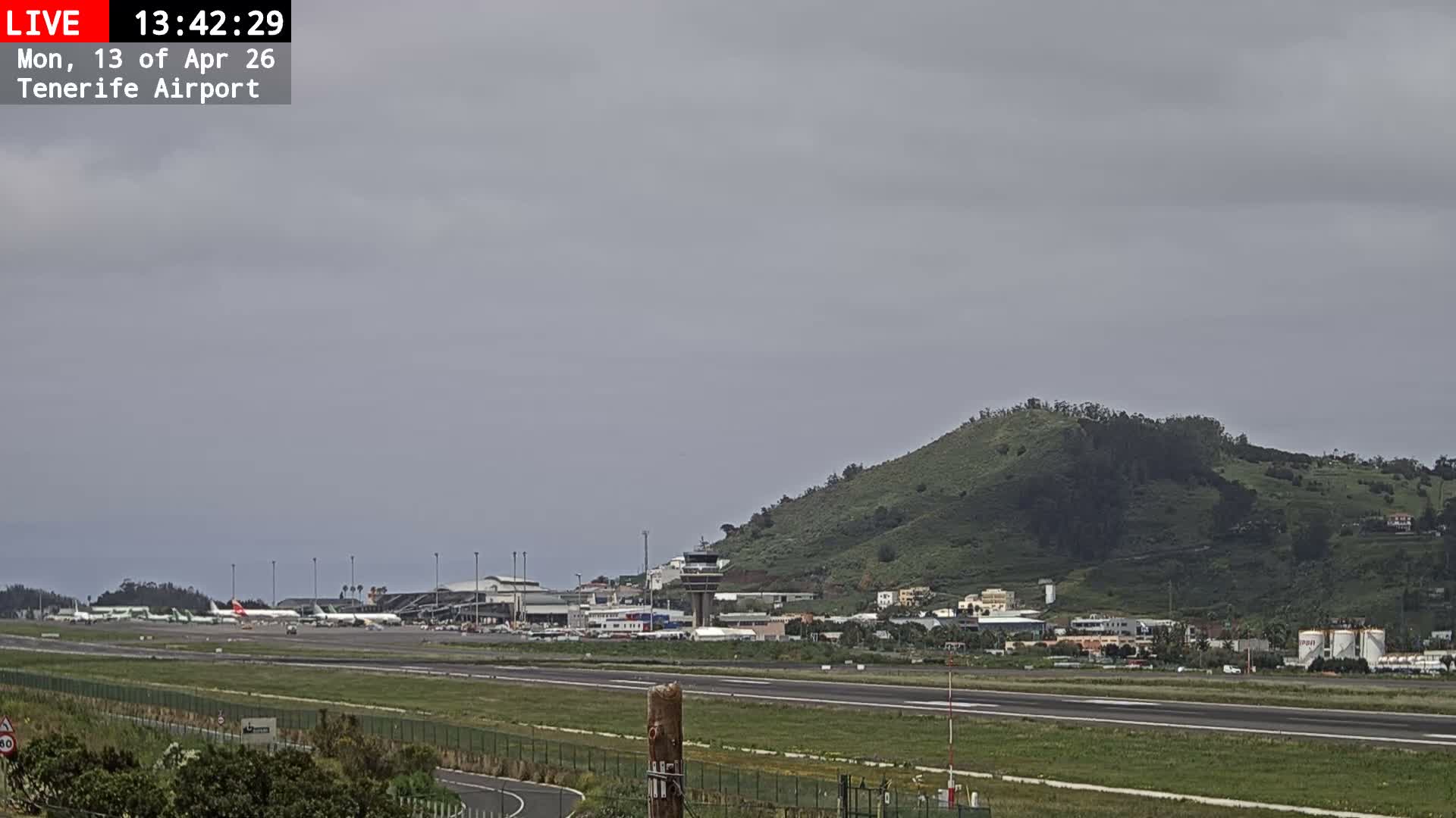 An airport runway and tarmac, featuring multiple airplanes, terminal buildings, and a control tower, are set against a backdrop of a large, tree-covered mountain under a uniformly overcast sky.