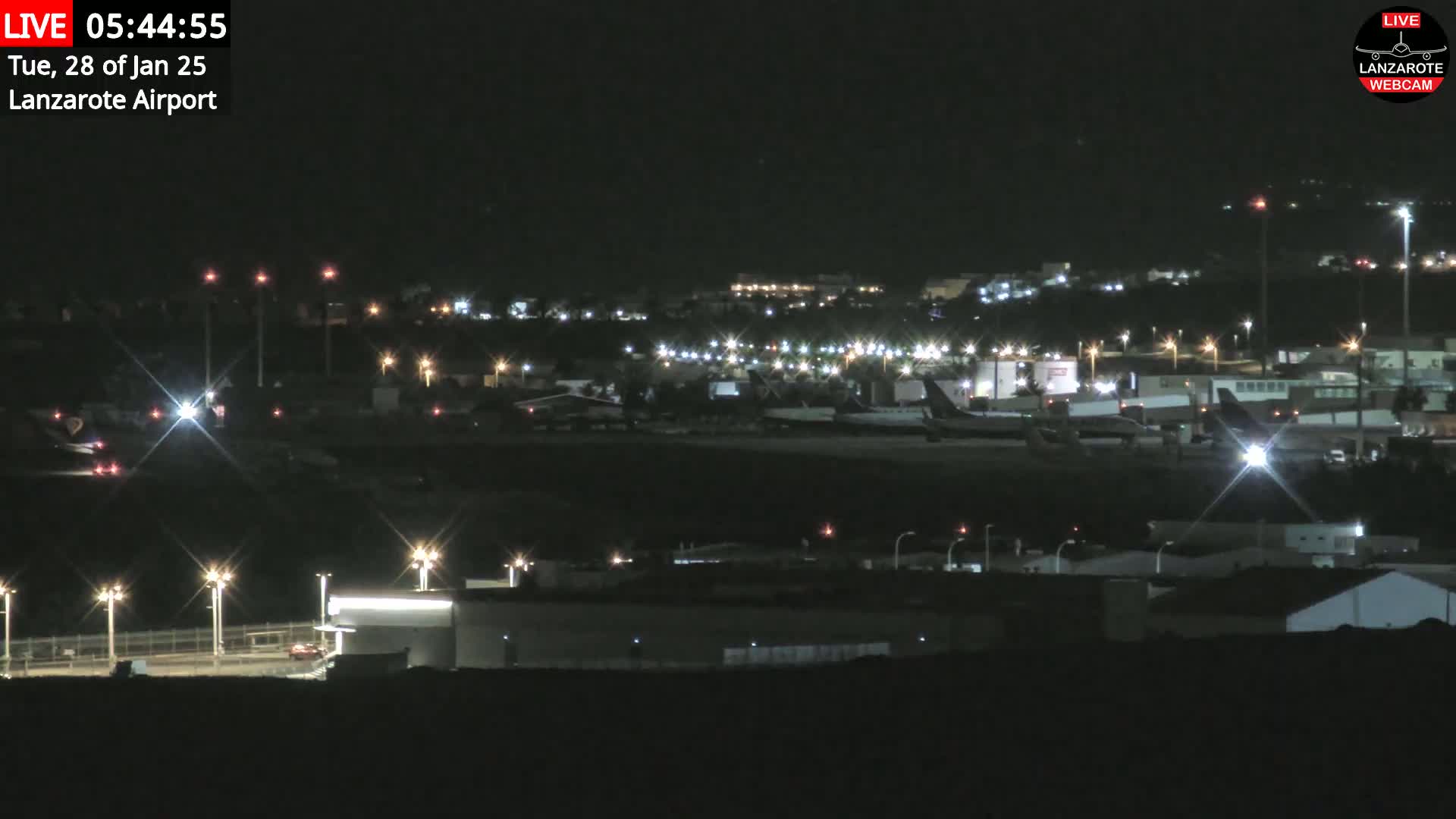 A nighttime, long shot of an airport tarmac with several airplanes and surrounding buildings illuminated by artificial lights.