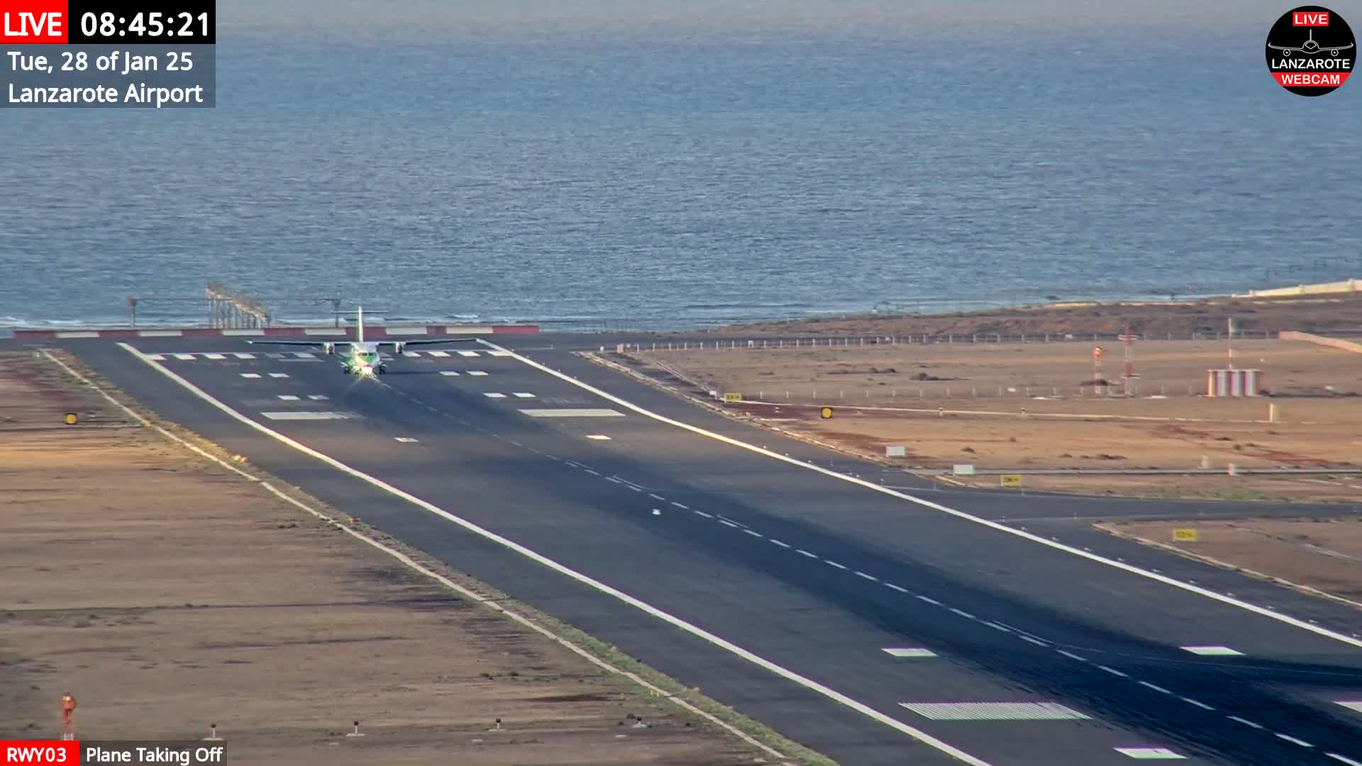 A small plane is taking off from a runway next to a calm ocean under a mostly clear sky.