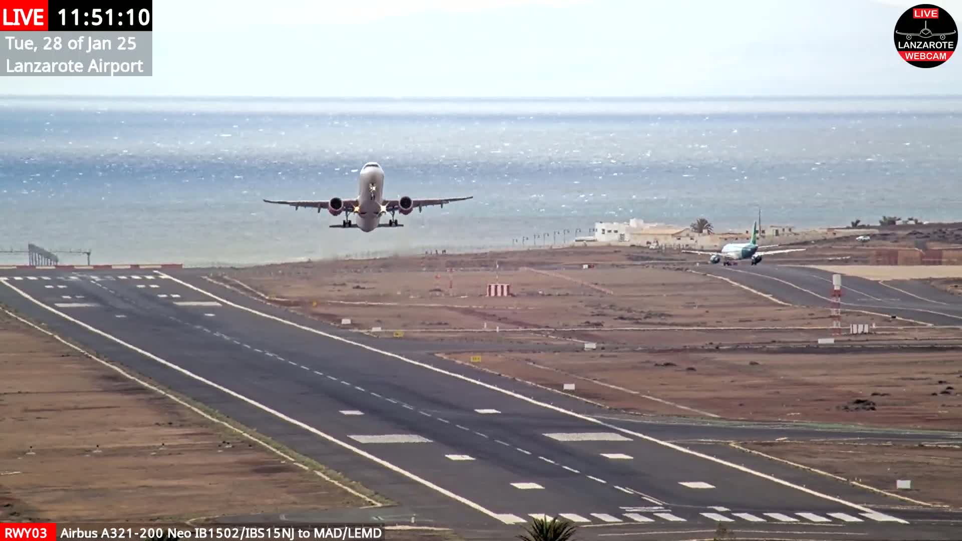 A passenger airplane takes off from an airport runway near a calm ocean under a mostly clear sky.