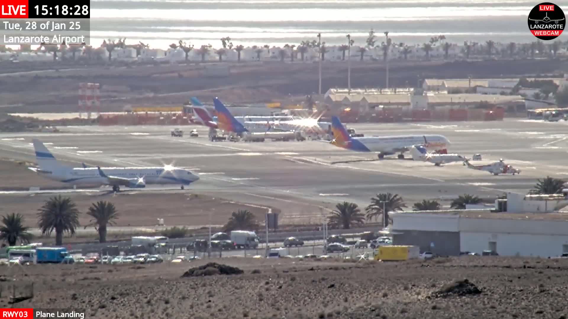 A bright, sunny day at an airport shows several airplanes on the tarmac, with one airplane landing on a runway, and palm trees and buildings visible in the background.