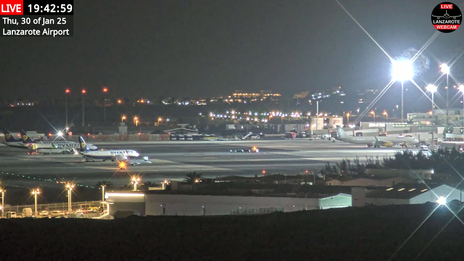 At night, several airplanes are parked on a brightly lit airport tarmac surrounded by buildings and vegetation.