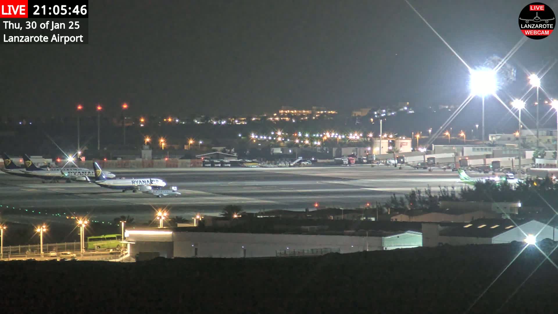 At night, several airplanes are parked on a brightly lit airport tarmac, with the surrounding area also illuminated.