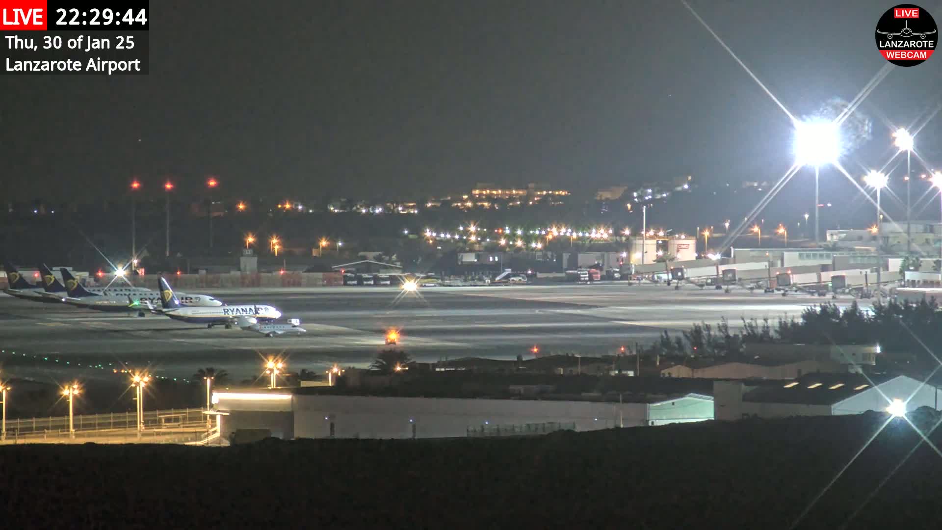 A nighttime view of an airport tarmac with several airplanes parked, illuminated by bright lights under a clear, dark sky.