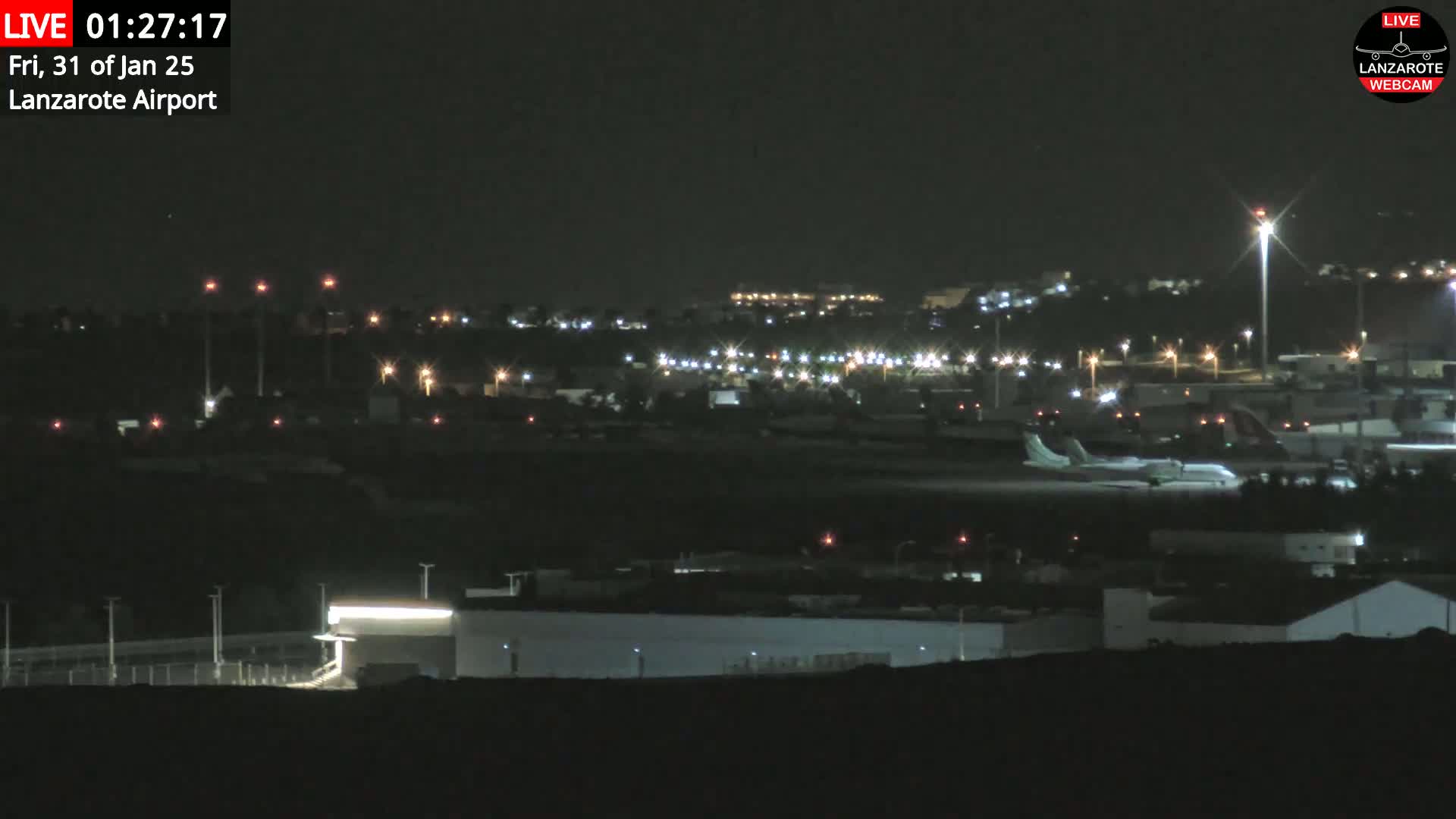 A nighttime, long shot of an airport with a plane on the tarmac, surrounded by dimly lit buildings and lights.