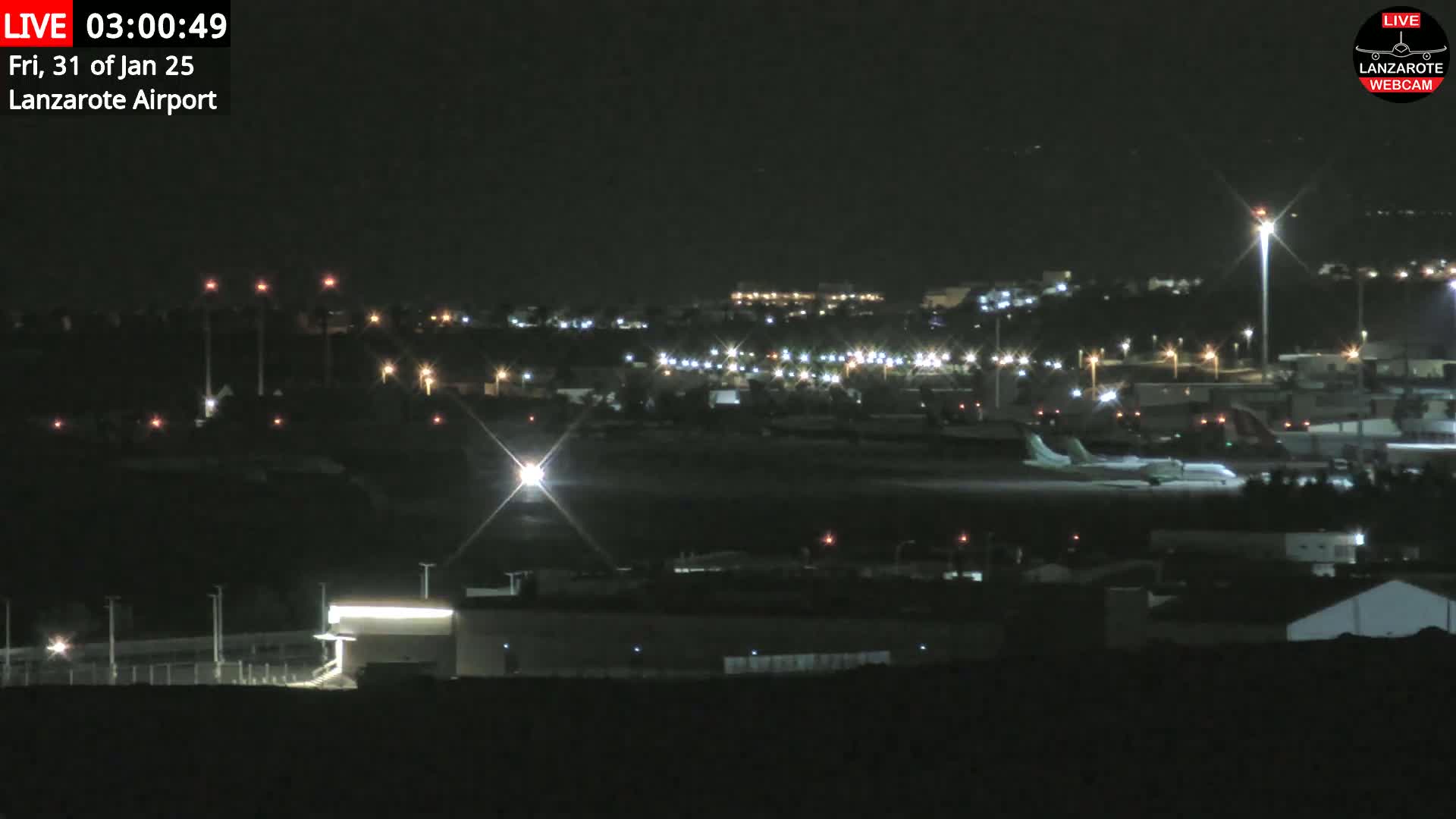 A nighttime view of an airport tarmac with a plane and various lights illuminating the surrounding buildings and landscape under a clear, dark sky.