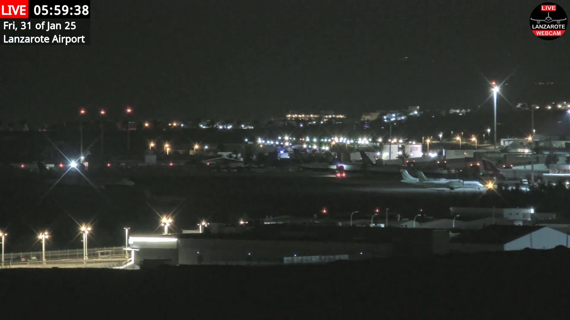 A nighttime long shot of an airport tarmac with a plane parked and several buildings illuminated by lights under a clear, dark sky.