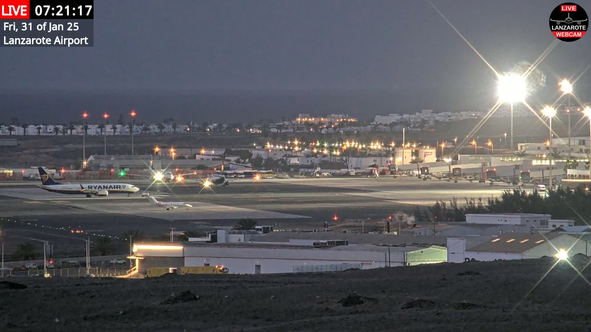 A nighttime view of an airport runway with several planes, including a Ryanair plane, under a dark, twilight sky.