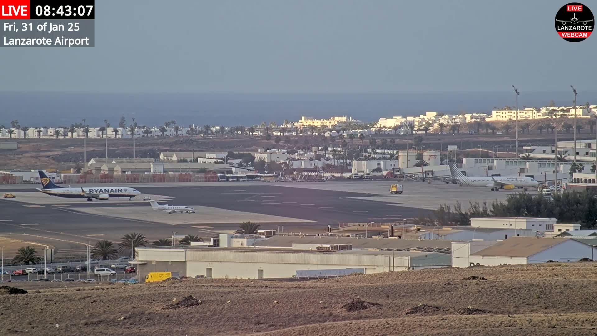 A mostly clear day at an airport shows several airplanes on the tarmac, with buildings and a body of water visible in the distance.