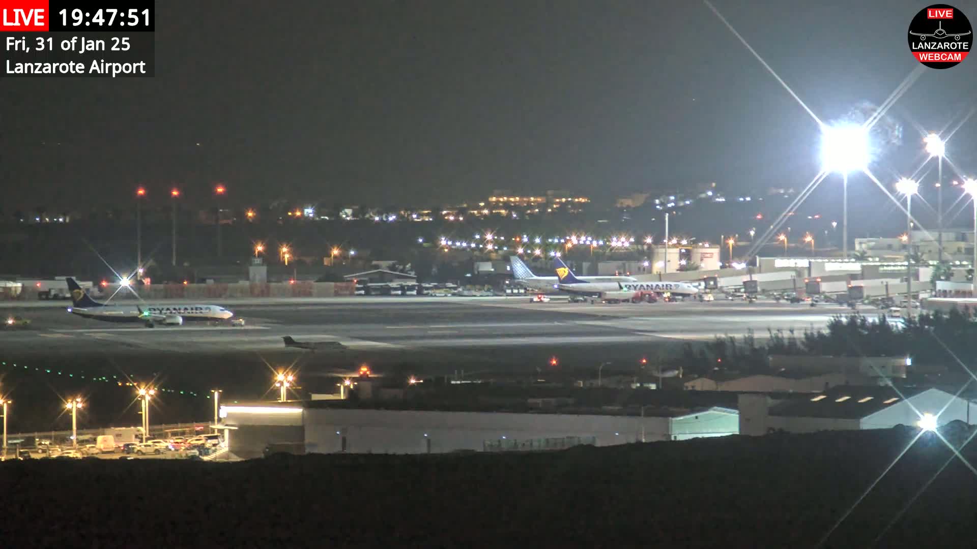 A nighttime view of an airport tarmac with two airplanes and various airport buildings illuminated under bright lights.