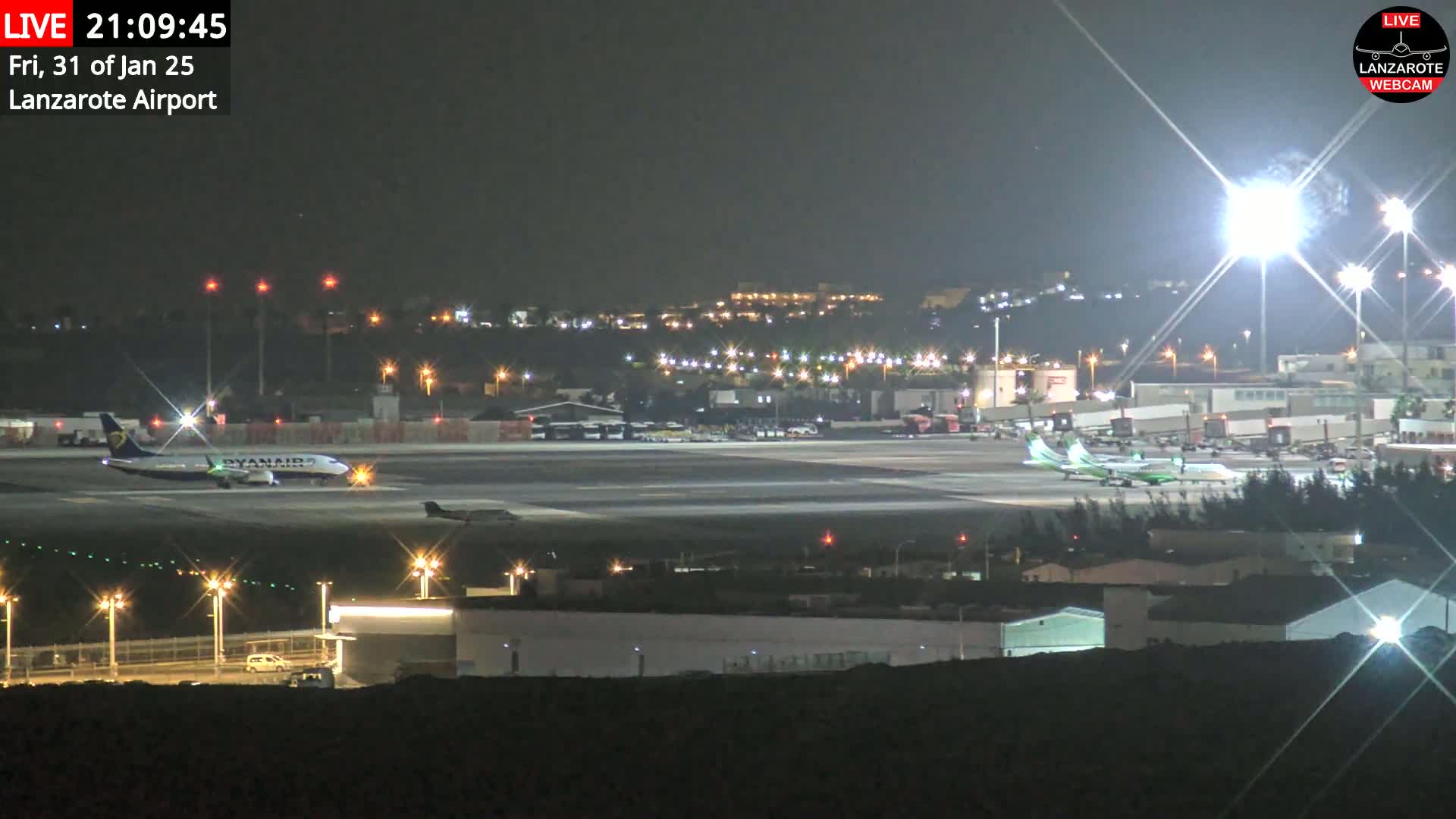A nighttime view of an airport tarmac with several airplanes, brightly lit buildings, and vehicles, under clear skies.