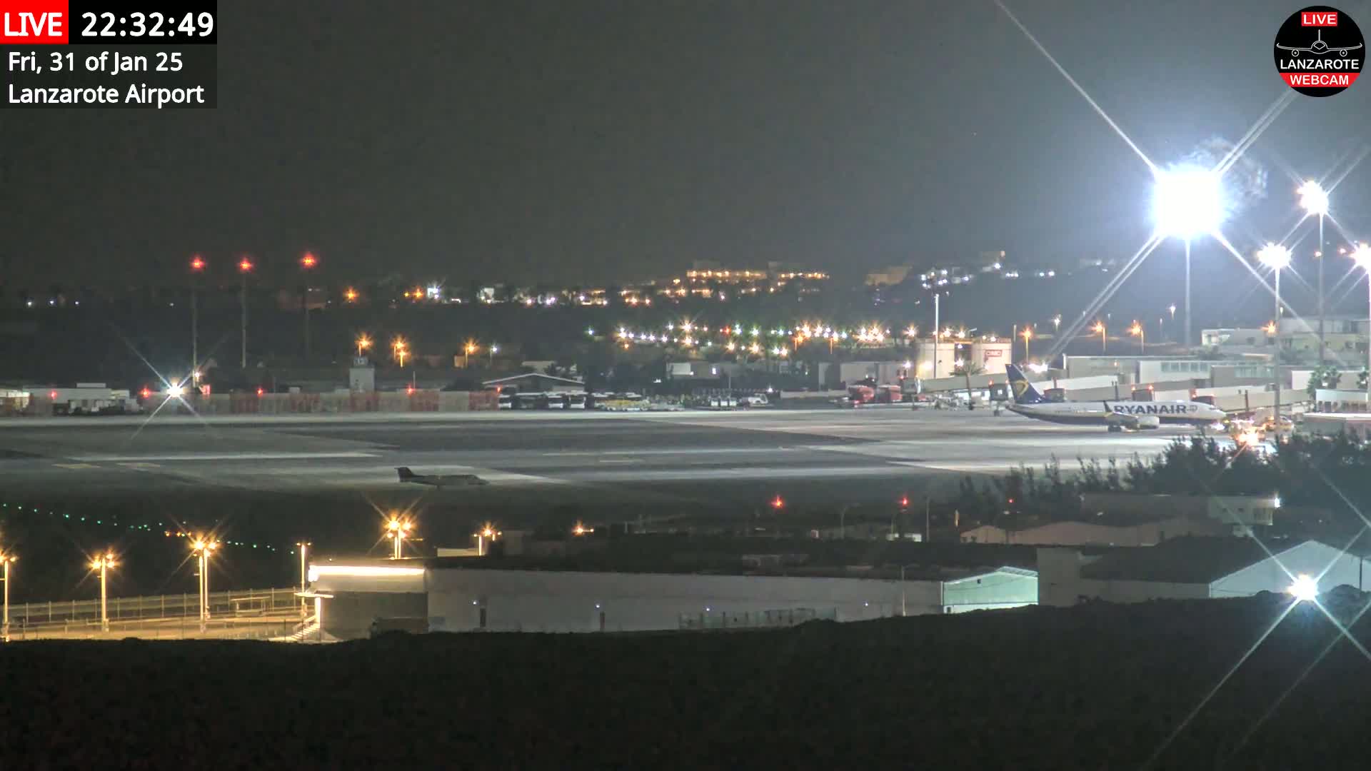 A nighttime, brightly lit airport tarmac shows several airplanes and airport vehicles, with city lights visible in the background under a clear, dark sky.