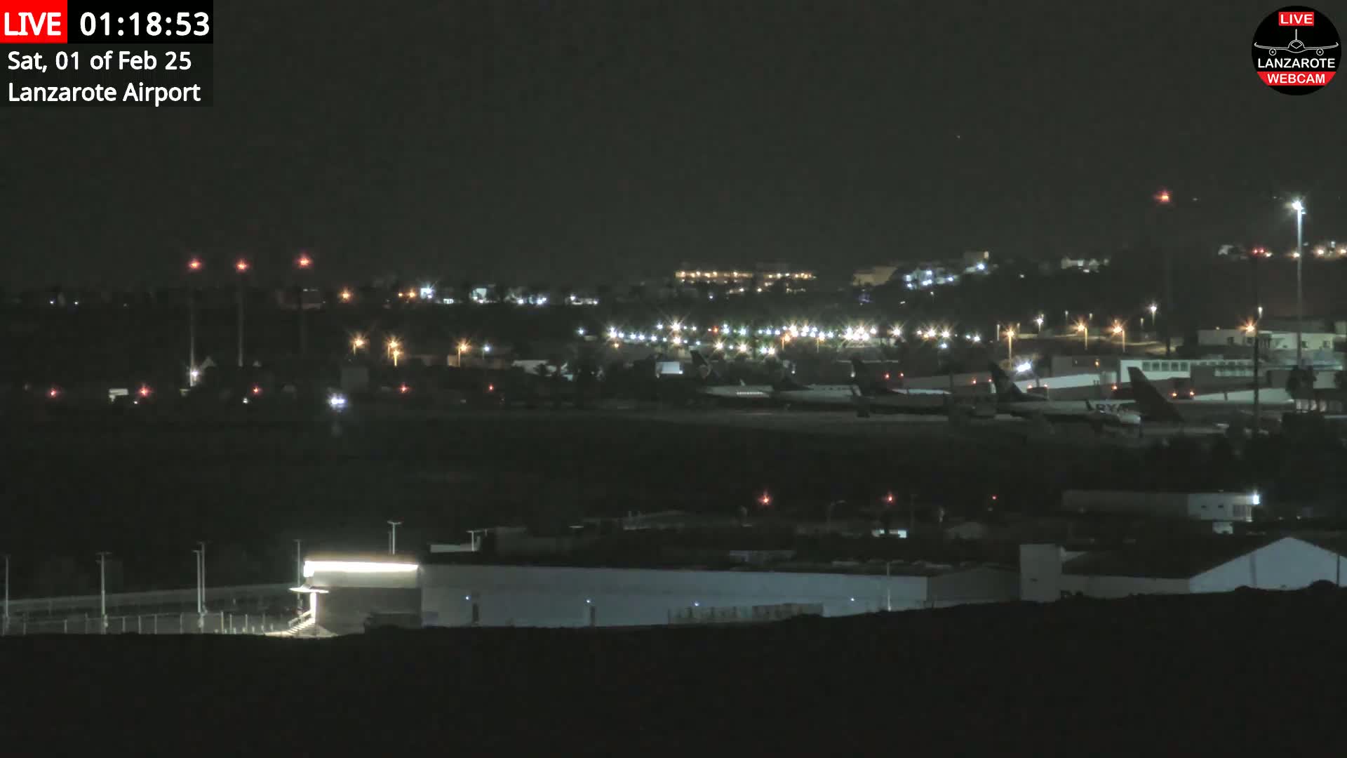 A nighttime long shot of an airport tarmac with several airplanes parked and illuminated by various lights under a clear, dark sky.
