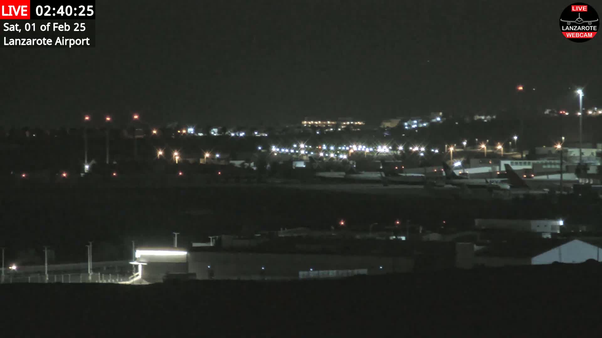 A nighttime long shot of an airport with numerous parked airplanes and illuminated buildings under a clear, dark sky.