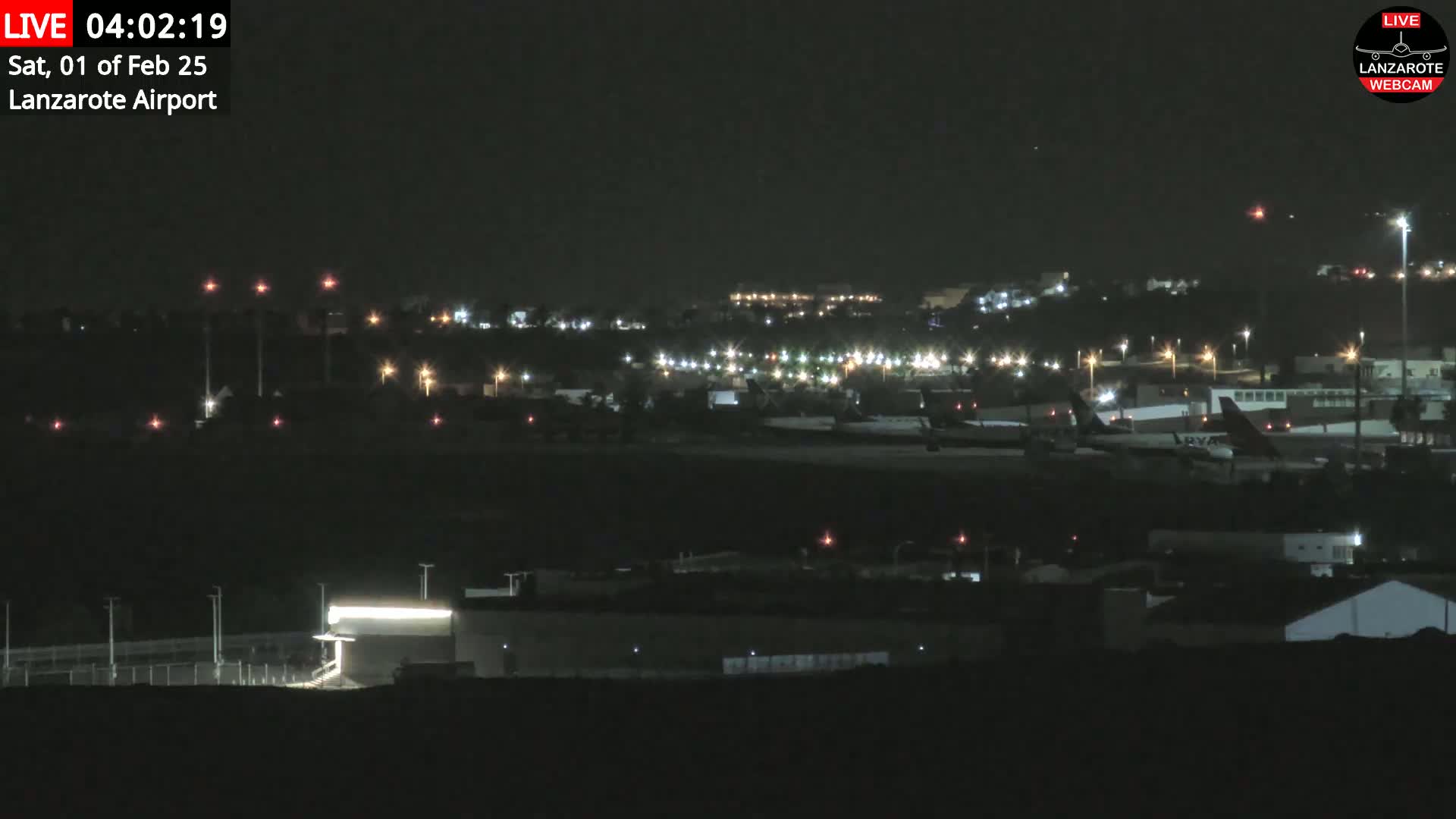 A nighttime long shot of an airport with several airplanes on the tarmac, illuminated by runway lights and surrounding building lights under a clear, dark sky.