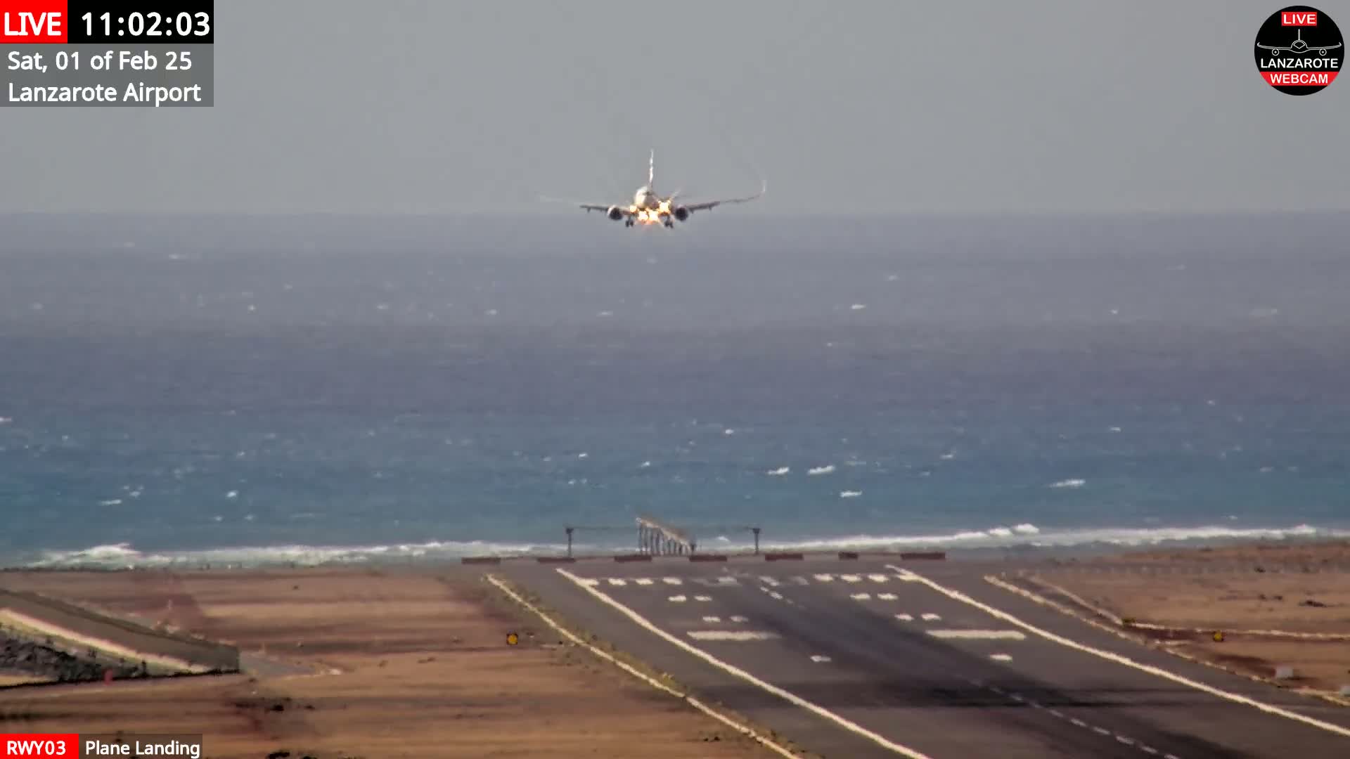A passenger plane is landing on a runway that borders a choppy ocean under an overcast sky.