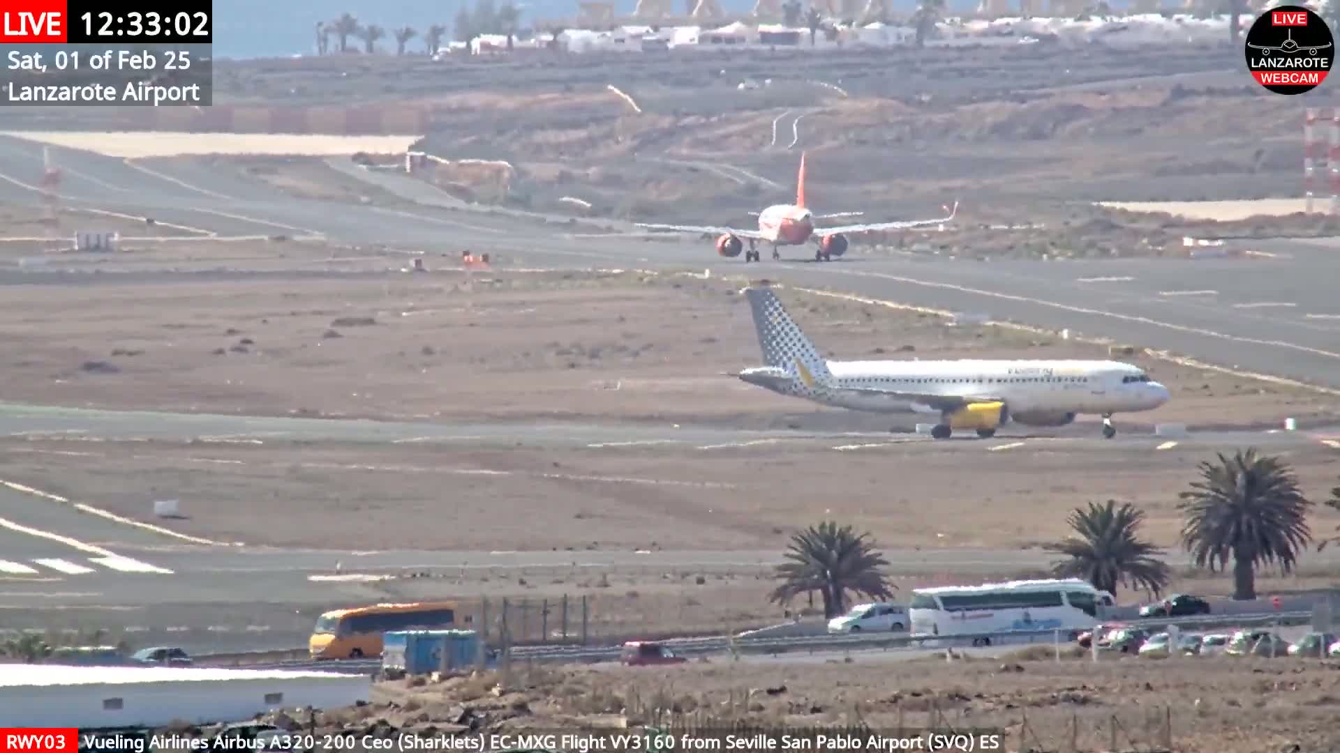 Two airplanes are on an airport tarmac under sunny conditions; one is taxiing and the other is near the end of a runway.