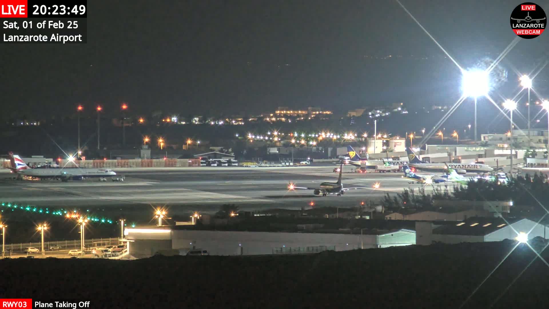 A nighttime airport scene shows several airplanes on the tarmac, with one plane taking off under bright lights.