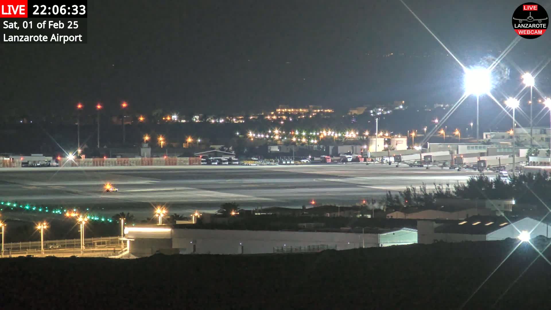 A brightly lit airport tarmac at night, with several planes visible near the terminal buildings, and surrounding city lights in the distance under a clear, dark sky.