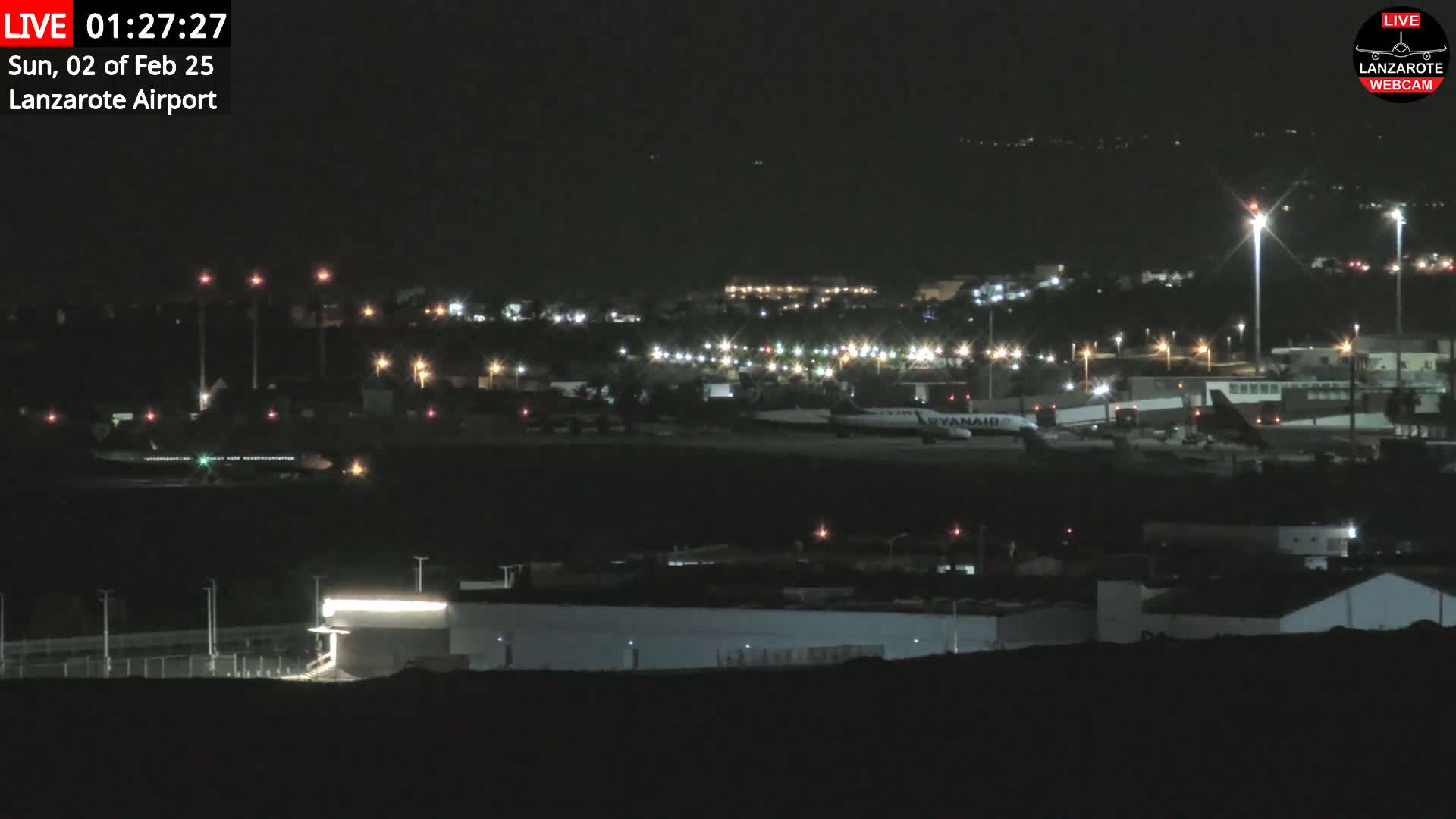 A nighttime view of an airport tarmac with airplanes and illuminated buildings under a clear, dark sky.