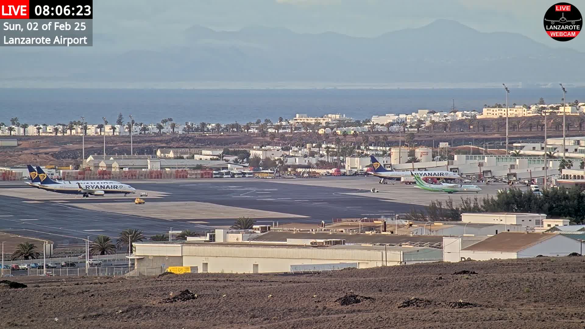 Two Ryanair airplanes are parked on an airport tarmac near buildings and a body of water, under a mostly clear sky with distant mountains visible.