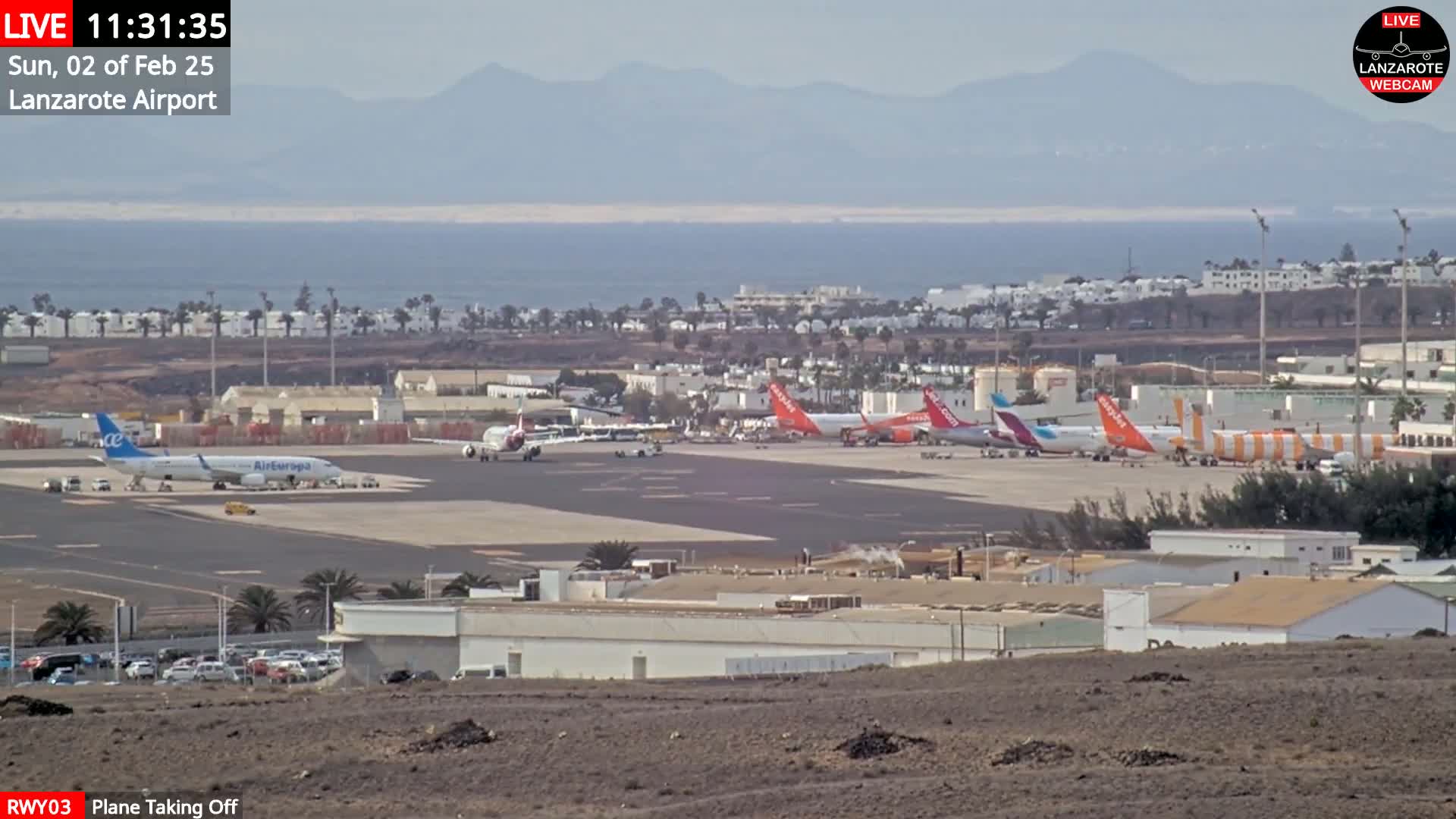 Several airplanes are parked on an airport tarmac near a coastline under hazy, sunny conditions with distant mountains visible.