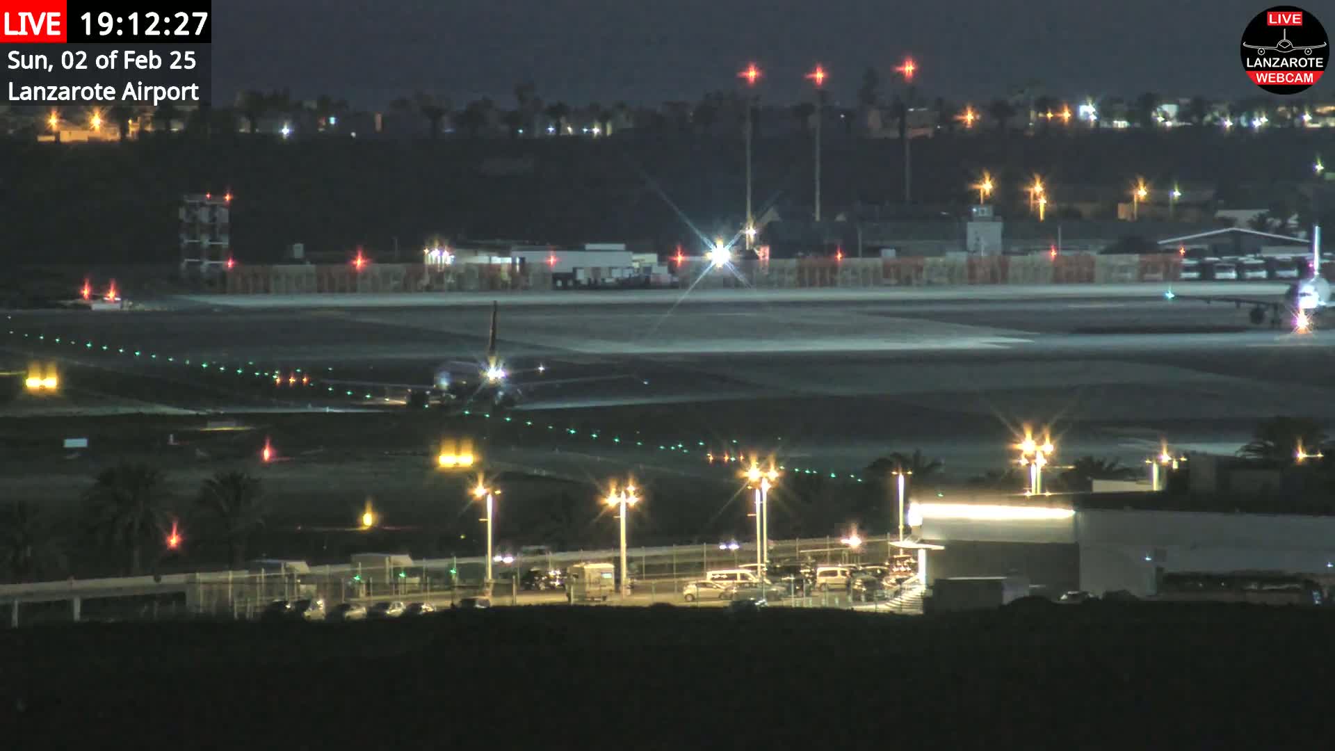 A nighttime view of an airport runway with an airplane taxiing, surrounded by illuminated buildings and vehicles.
