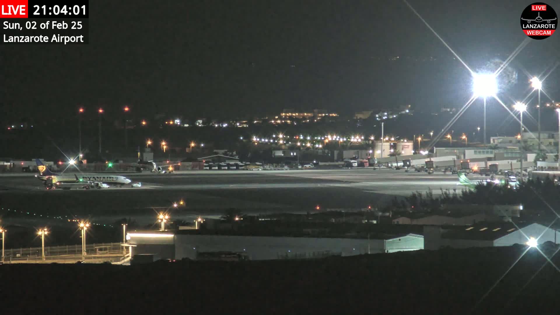 A nighttime view of an airport tarmac with several planes, ground support vehicles, and buildings illuminated by bright lights.
