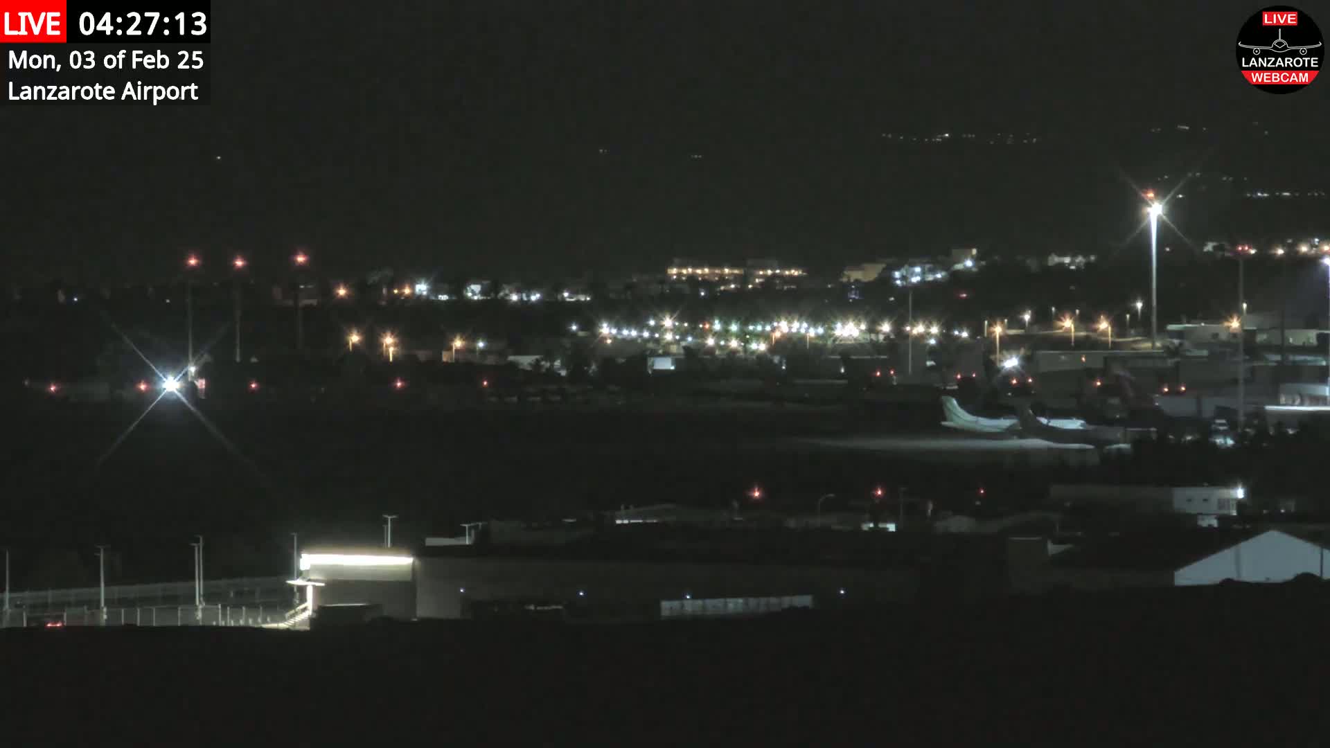 A nighttime, long shot of an airport tarmac with various buildings and lights illuminated under a clear, dark sky.