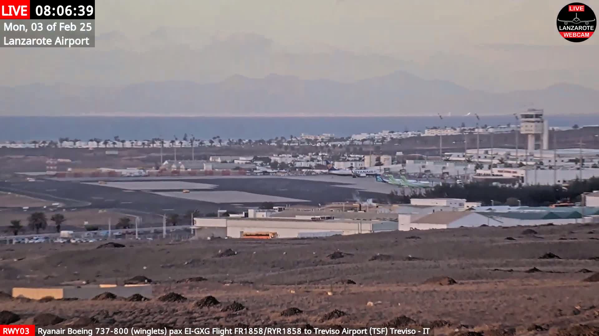 An airport with several airplanes on the tarmac is shown, with a calm ocean and distant mountains visible in the hazy background.