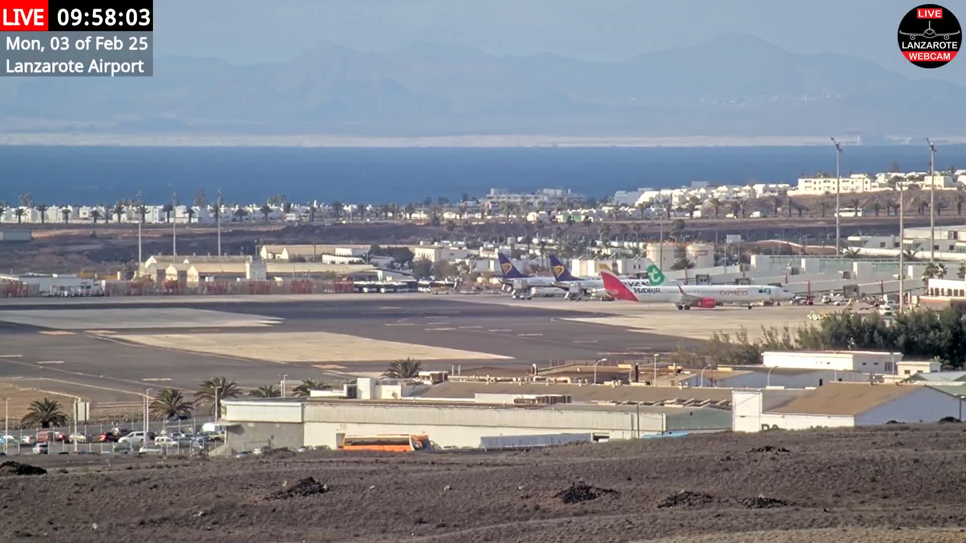 A sunny day at an airport shows several airplanes parked on the tarmac near terminal buildings, with a calm ocean in the background.