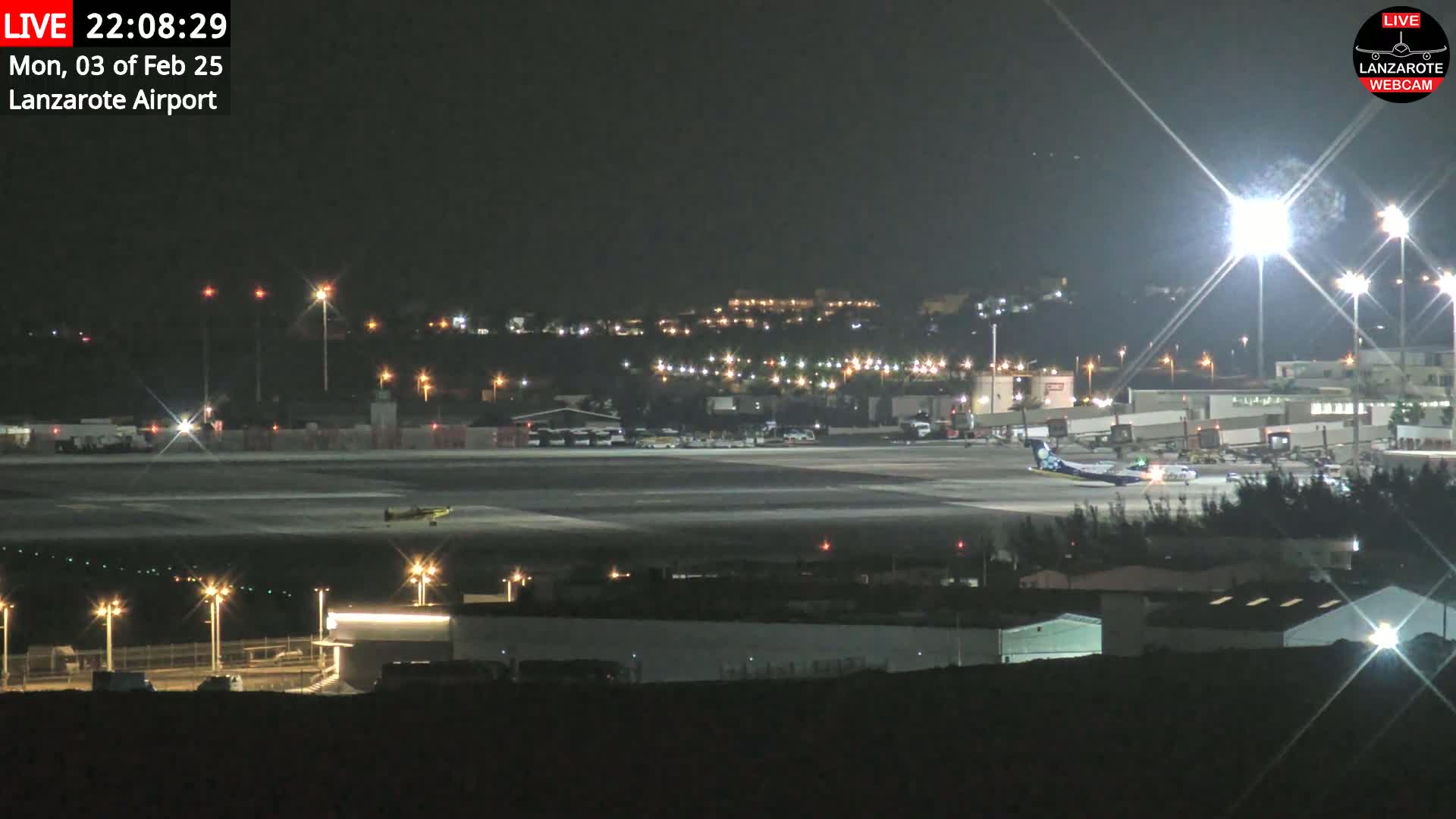 A nighttime view of an airport tarmac with several airplanes, ground vehicles, and brightly lit buildings, under clear skies.