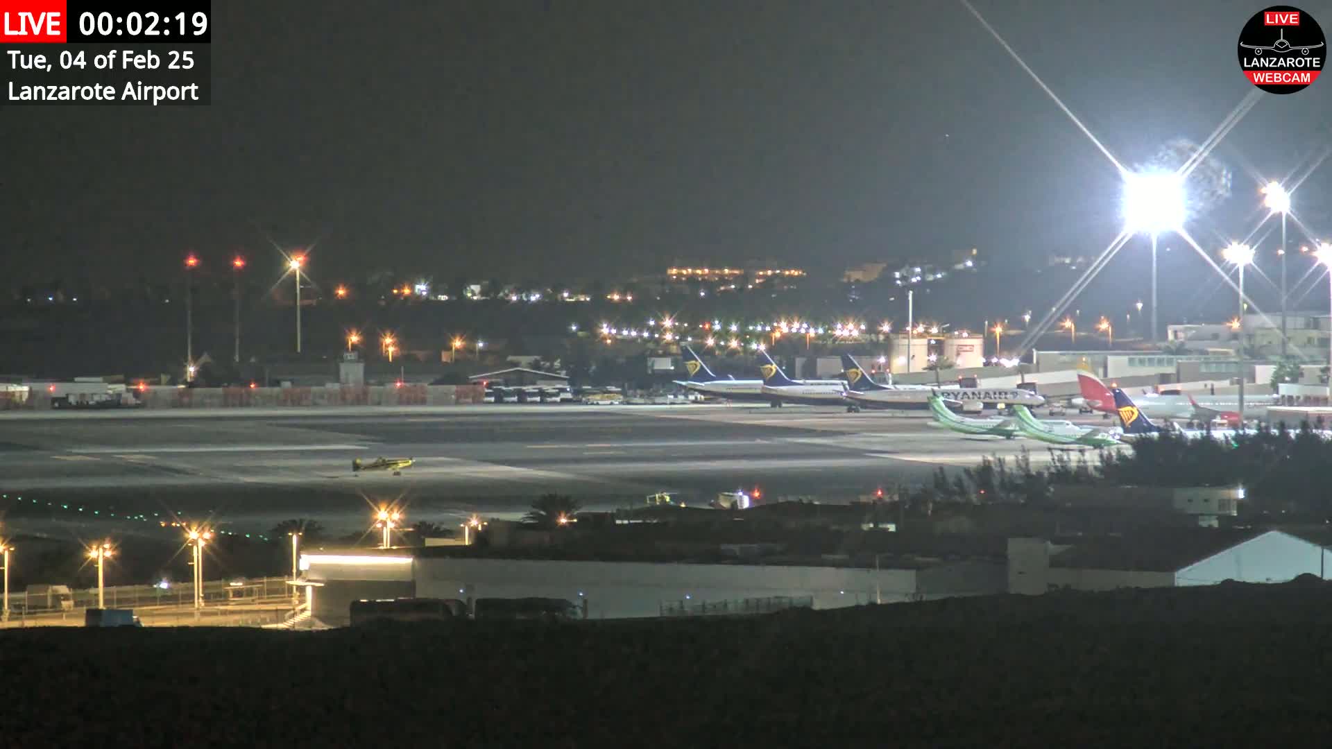 A nighttime view of a brightly lit airport tarmac with several airplanes parked, under a clear, dark sky.