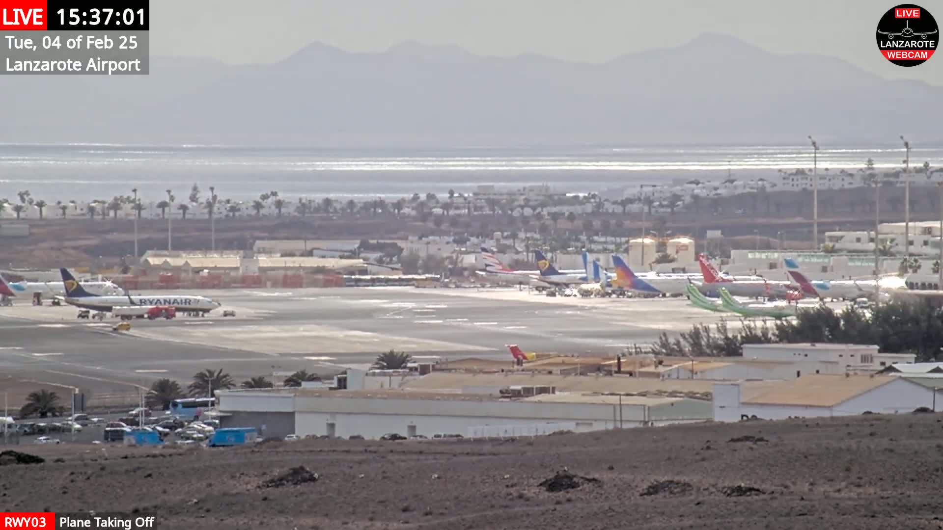 A Ryanair plane is taking off from an airport with several other planes parked on the tarmac, with a calm ocean and distant mountains visible in the background under a hazy sky.