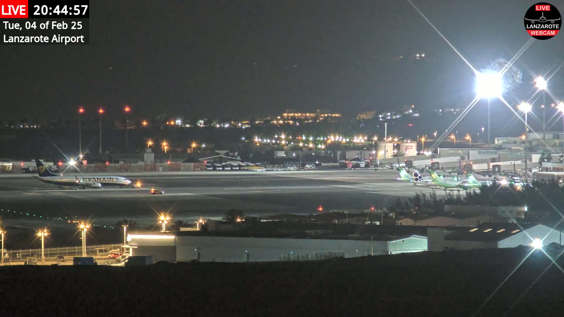 A brightly lit airport tarmac at night, featuring several airplanes, including a Ryanair plane, under clear skies.